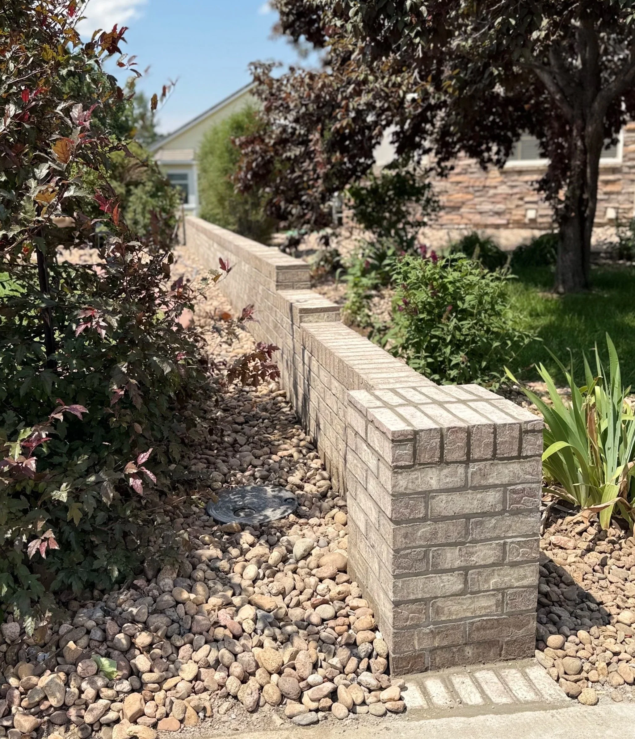 A landscaped garden area with a brick retaining wall, small decorative rocks, green plants, and trees in front of a house with a stone exterior and a sloped roof.