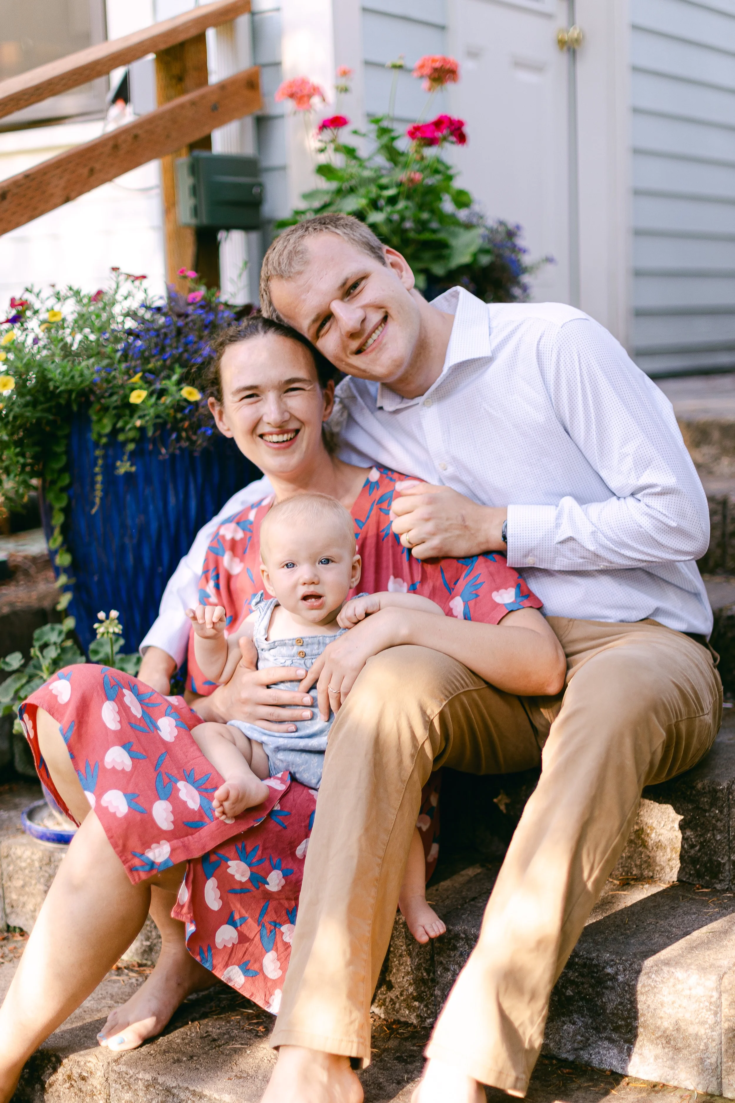 A smiling family of three sitting on steps outside near flowers and a light blue house, with a woman, man, and baby girl.
