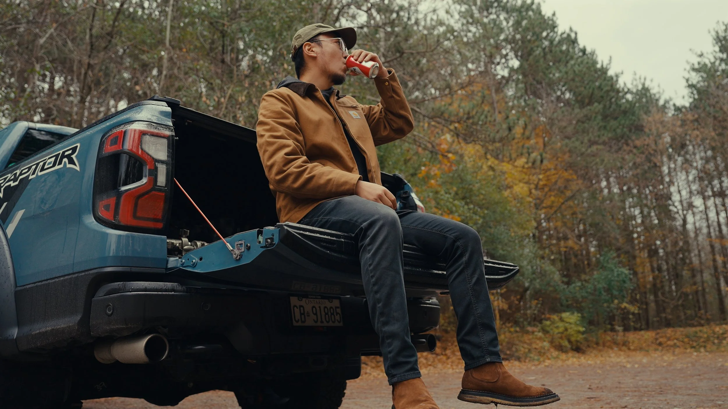 A man sitting on the open tailgate of a blue Ford Raptor pickup truck in a wooded area, drinking from a can amid autumn scenery.