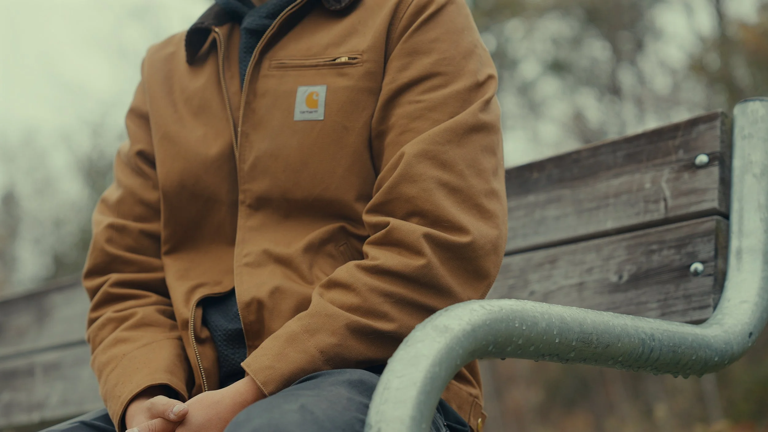 Close-up of a person in a brown Carhartt jacket sitting on a park bench with trees in the background.