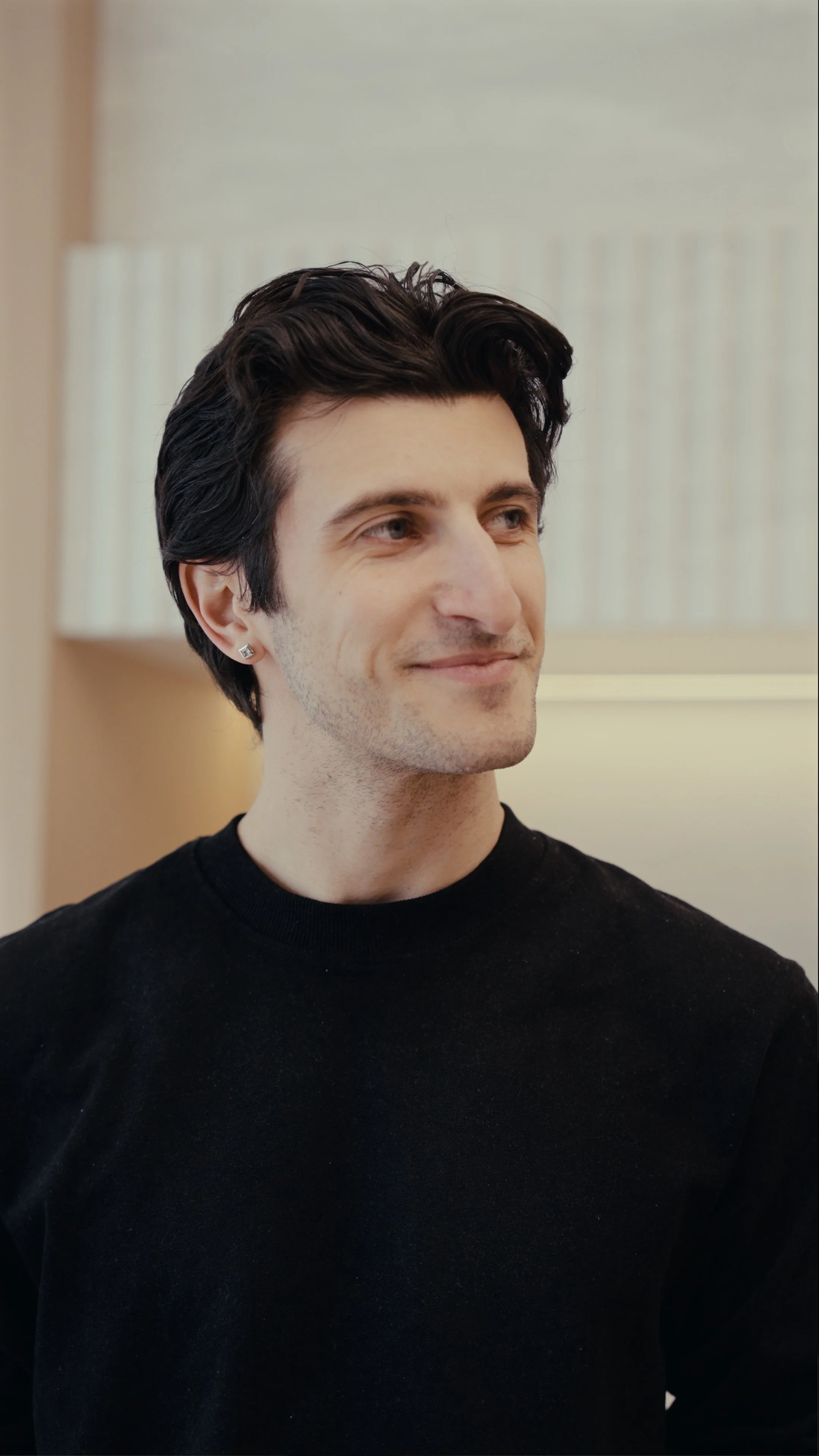 A young man with dark hair, wearing a black shirt and a small earring, smiling slightly in a softly lit room.