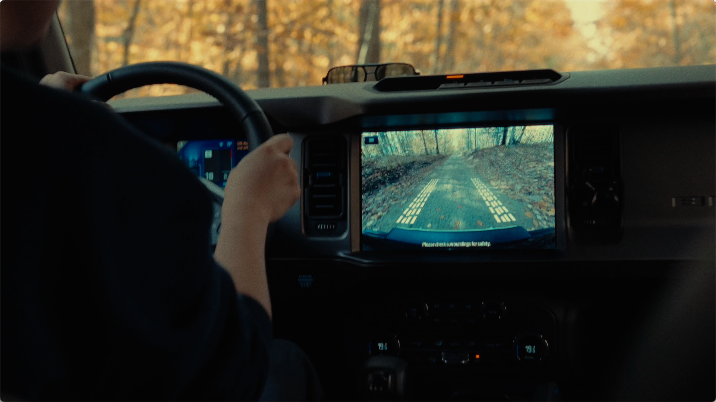 Inside a vehicle, a person is driving on a woodland trail with a view of the dashboard and backup camera display showing a forest path with fallen leaves.