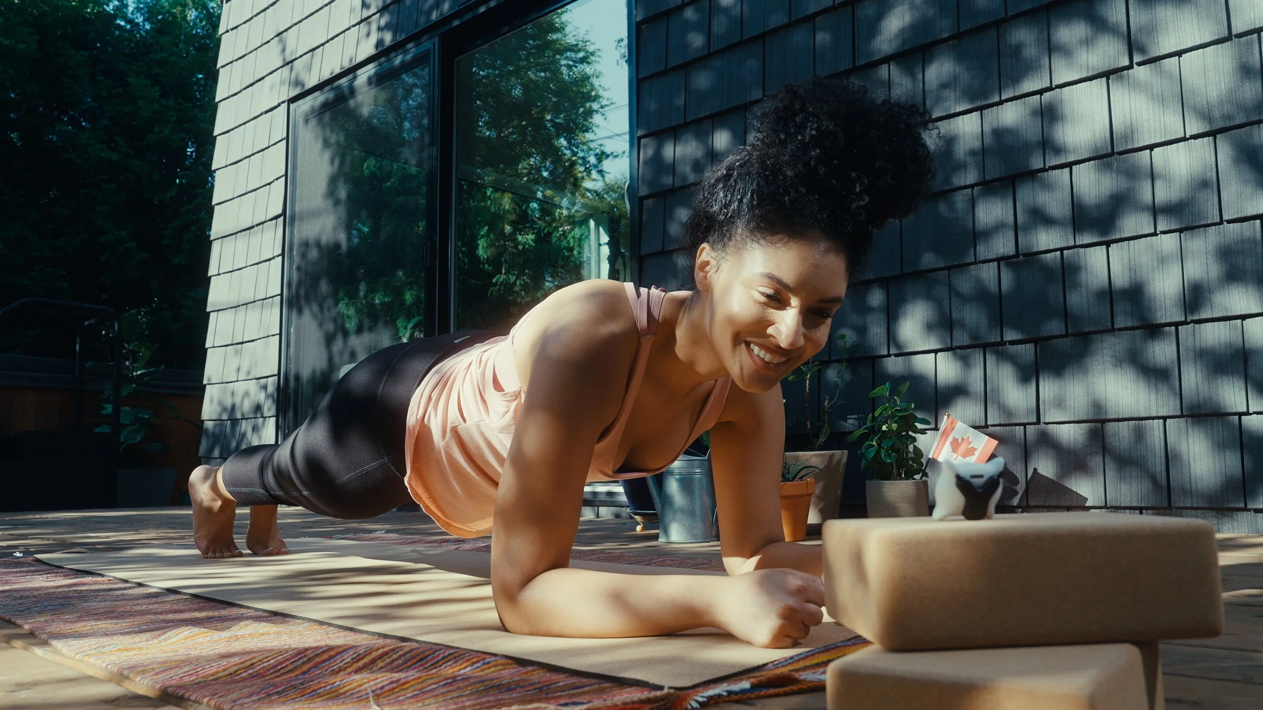 A woman doing a plank exercise outdoors on a yoga mat, smiling, with potted plants and a house wall in the background.