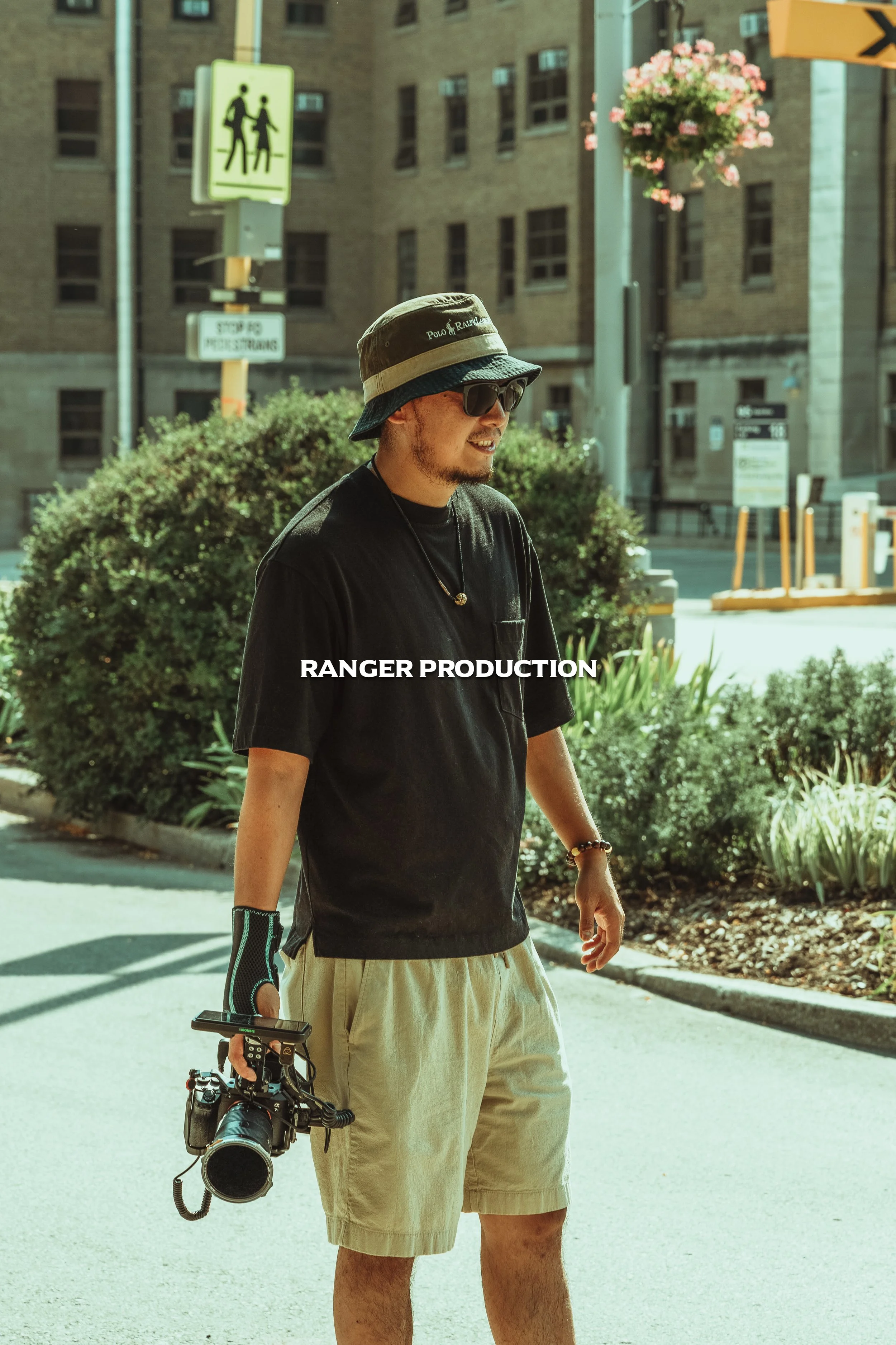 A man with a camera walking on a city street with bushes and buildings in the background.