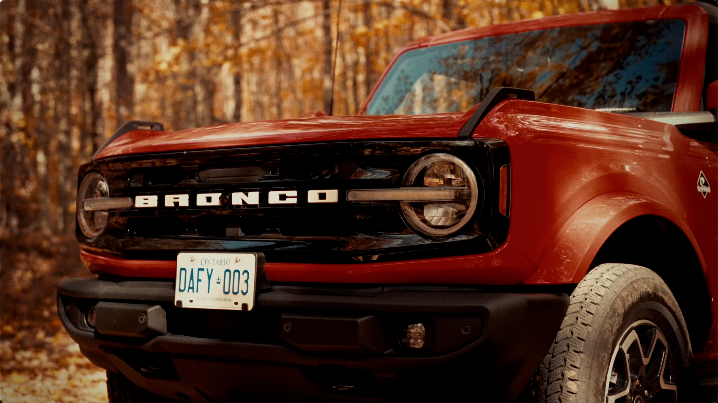 Close-up of a red Ford Bronco with black grille and Ontario license plate 'DAFY-003', parked on a dirt road with autumn trees in the background.