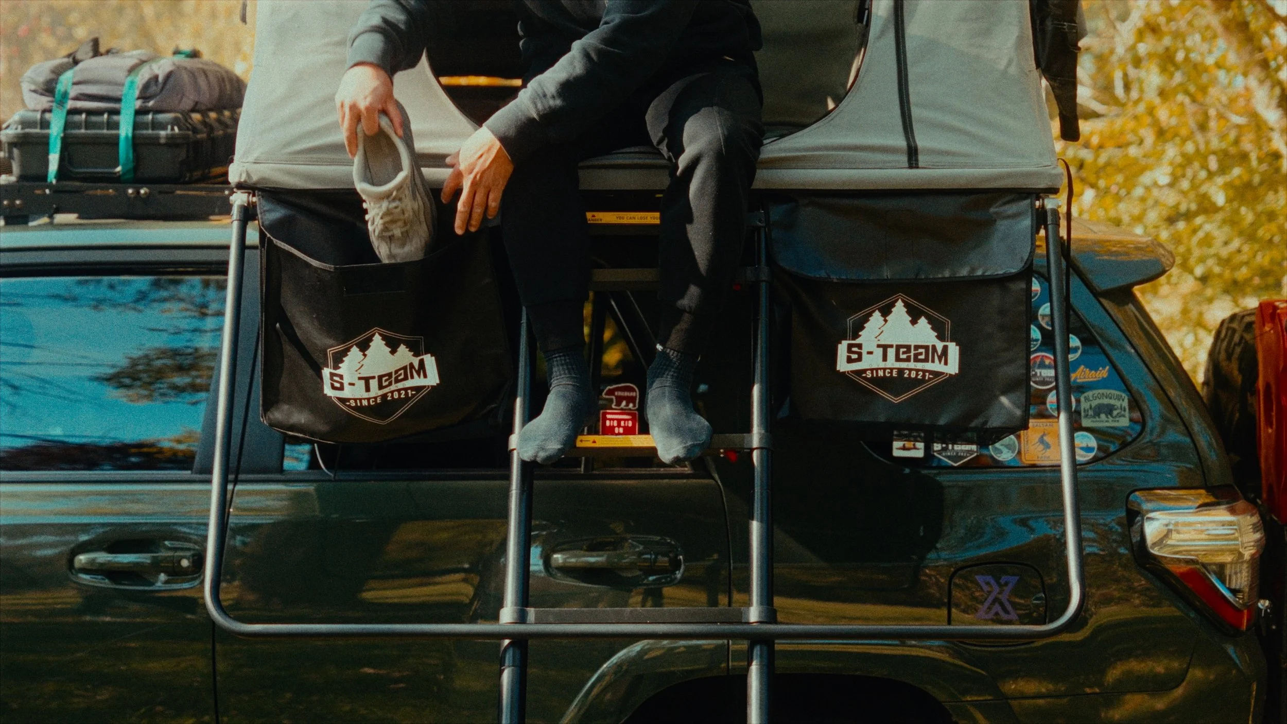 Person sitting in the back of a vehicle with a rooftop tent, holding a pair of shoes, surrounded by camping gear and stickers in an outdoor setting.