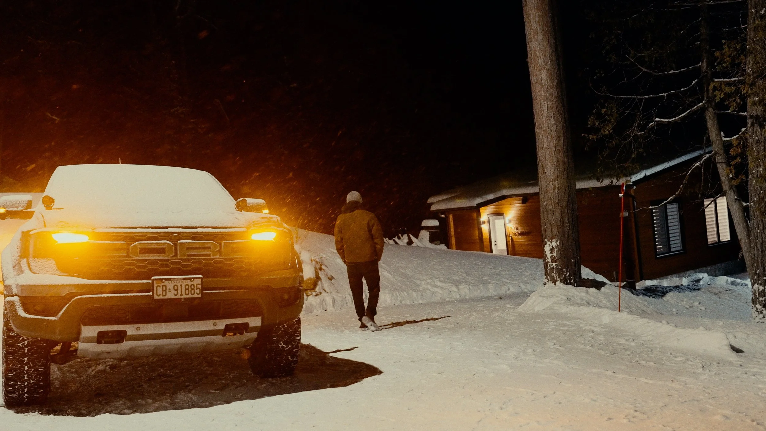 A person in winter clothing walking away from a snow-covered pickup truck with glowing headlights, outside a cabin at night in a snowy landscape.