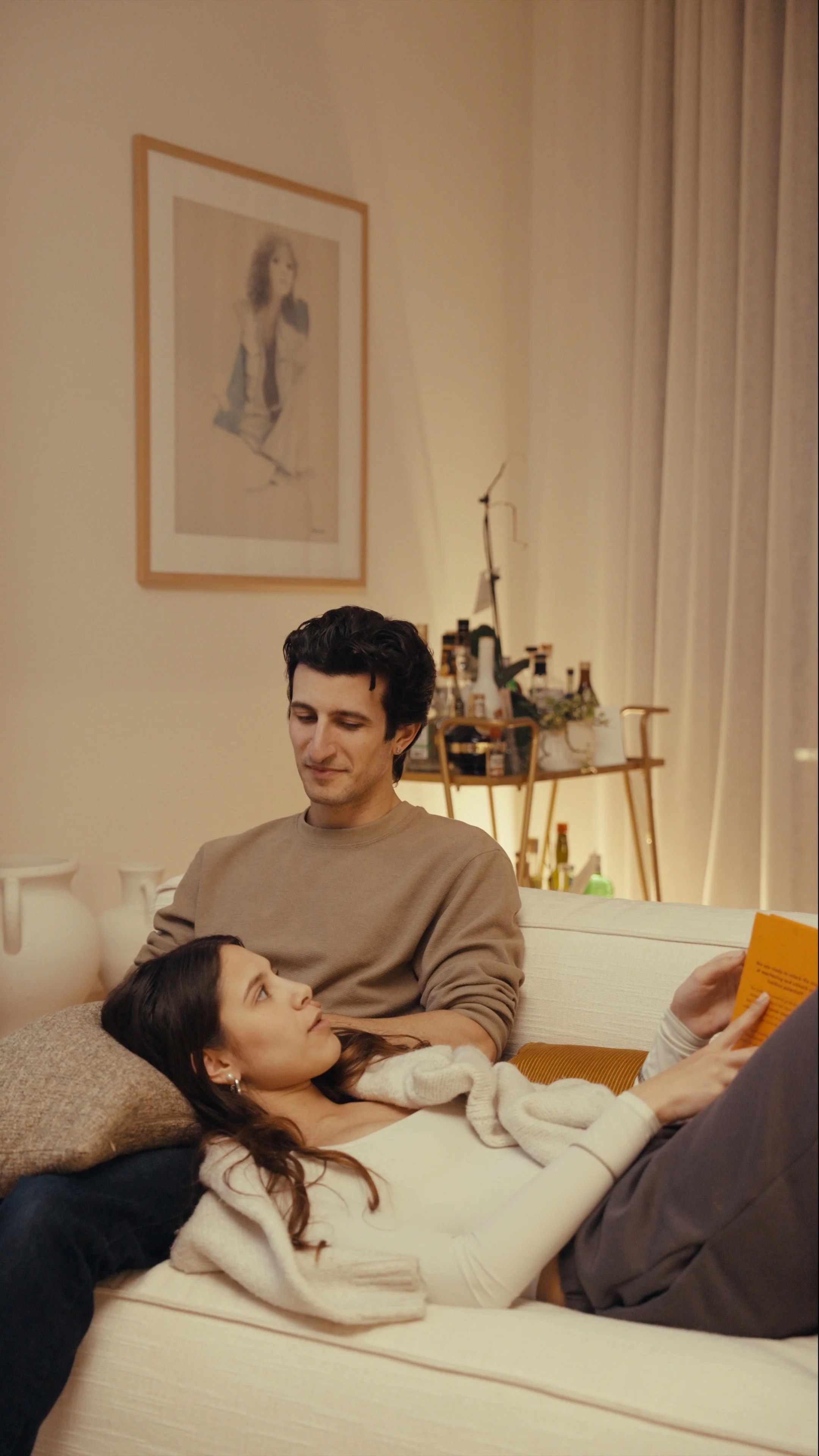 A couple sitting on a sofa in a well-lit living room, with the woman lying down and reading a book while the man looks at her. The background has a framed picture on the wall and a bar cart with bottles.
