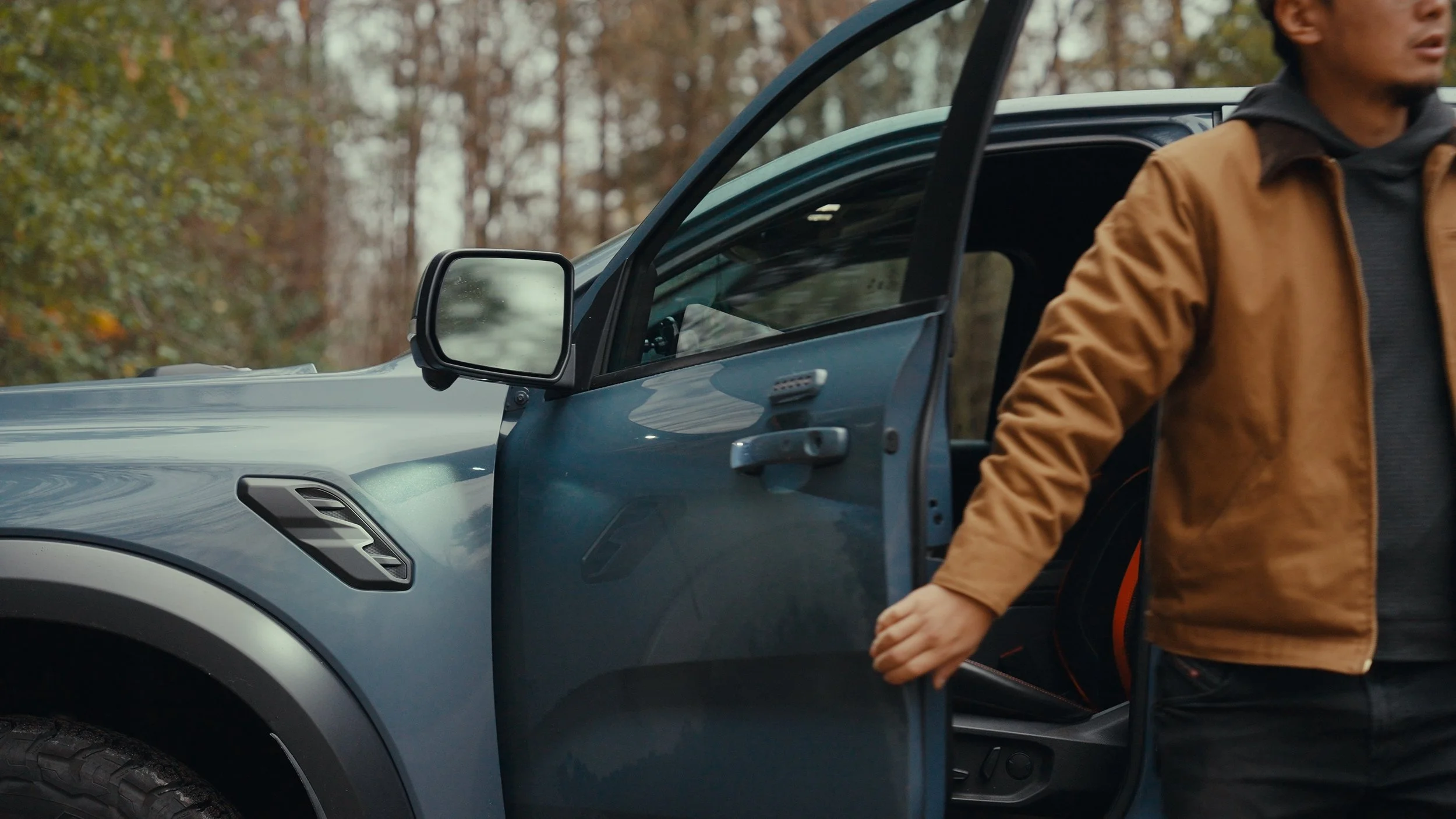 Person in brown jacket opening the driver’s side door of a blue vehicle in a wooded area.