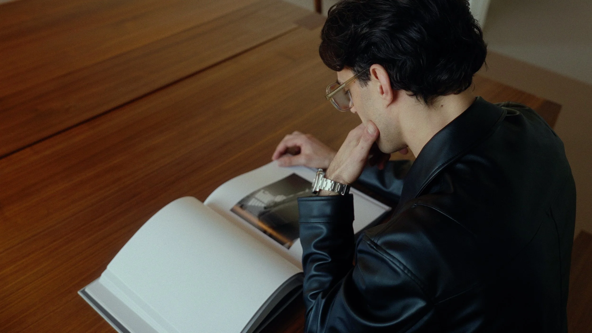 A man with curly black hair and glasses, dressed in a black leather jacket, is sitting at a wooden desk reading a book or magazine, with his right hand touching his chin.