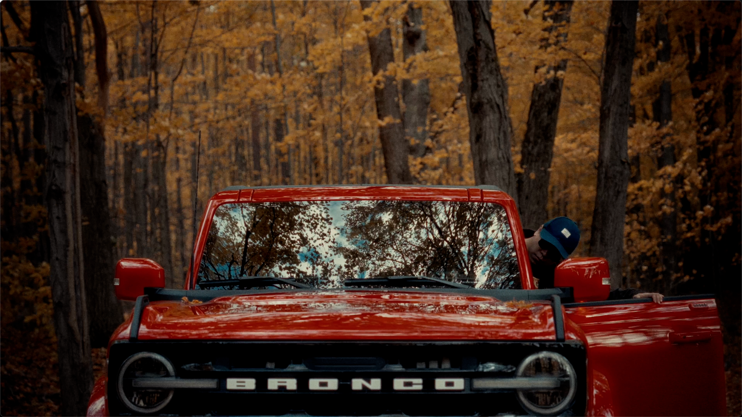 A person wearing a blue cap leaning out of the driver's side door of a bright red vintage car with the word 'BRONCO' on the front grille, parked in a forest with tall trees and autumn leaves.