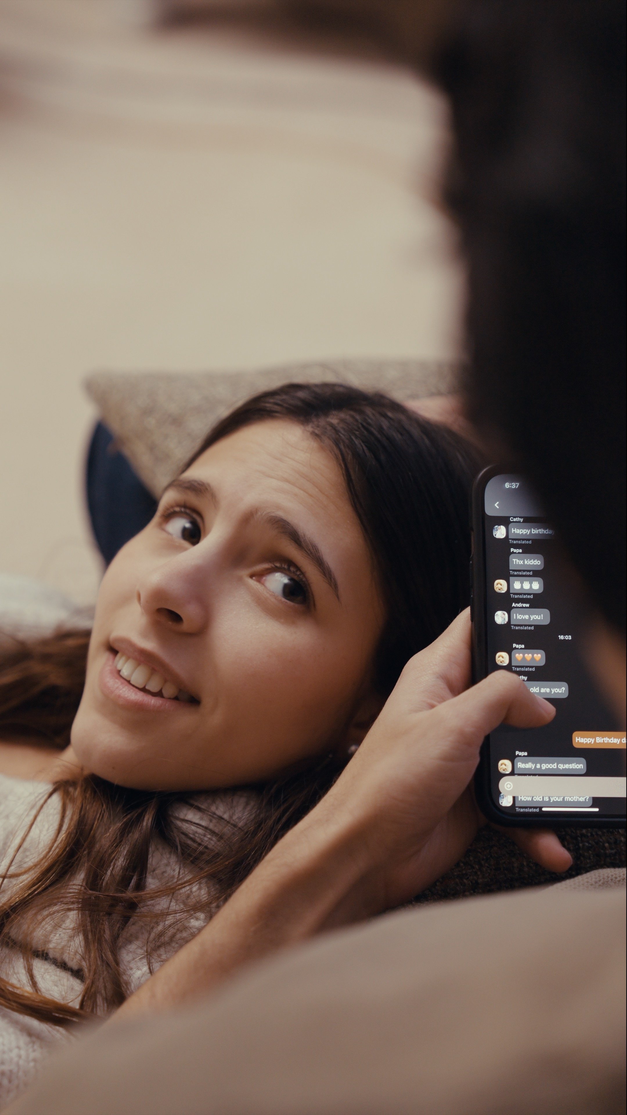 A young woman with long dark hair lying down on a couch, smiling and looking at a smartphone screen that displays a messaging conversation with birthday wishes and questions.