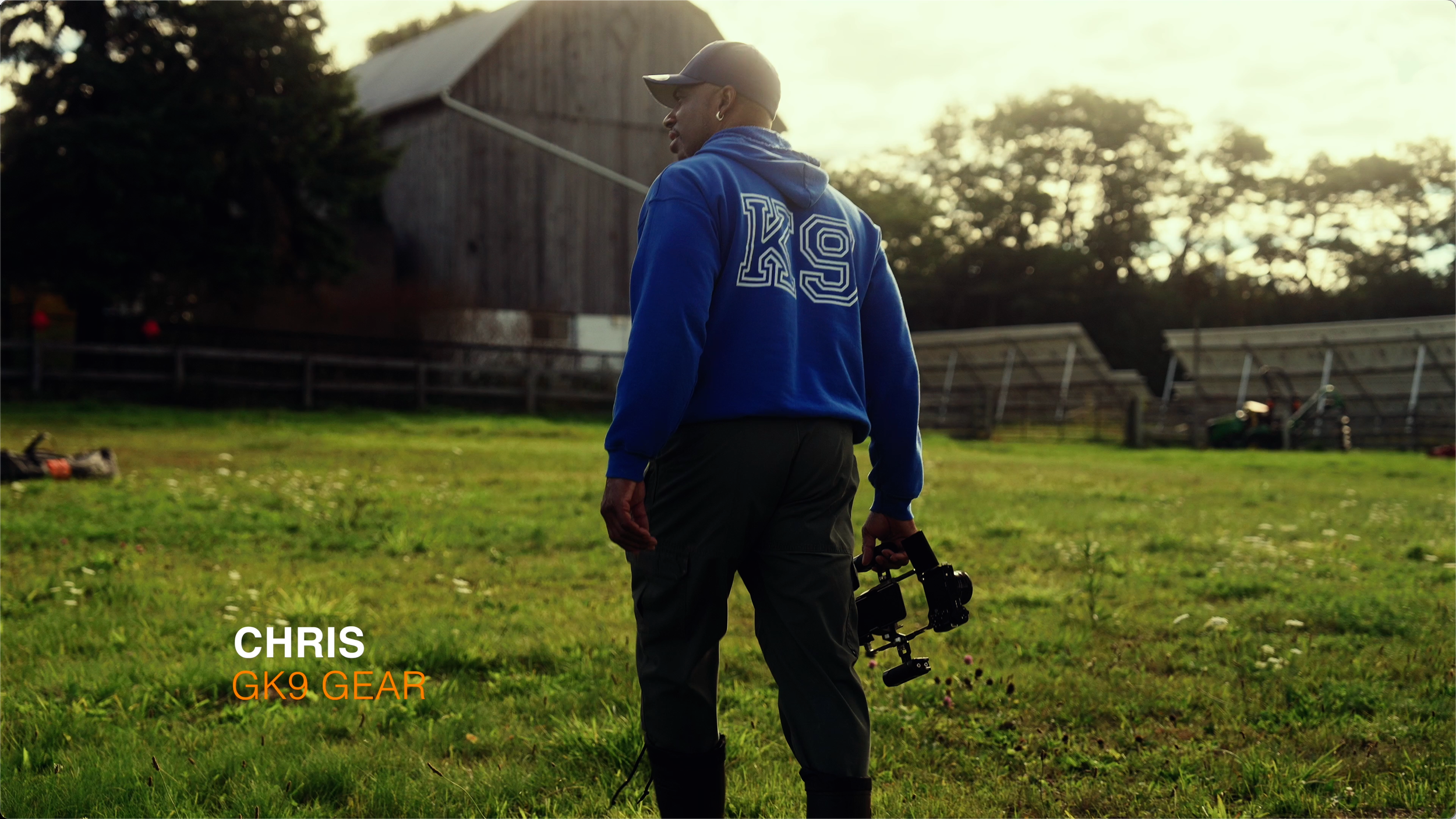 A man wearing a blue hoodie with 'GK9' on the back and a baseball cap, holding a camera stabilizer, standing on a grassy field during sunset.