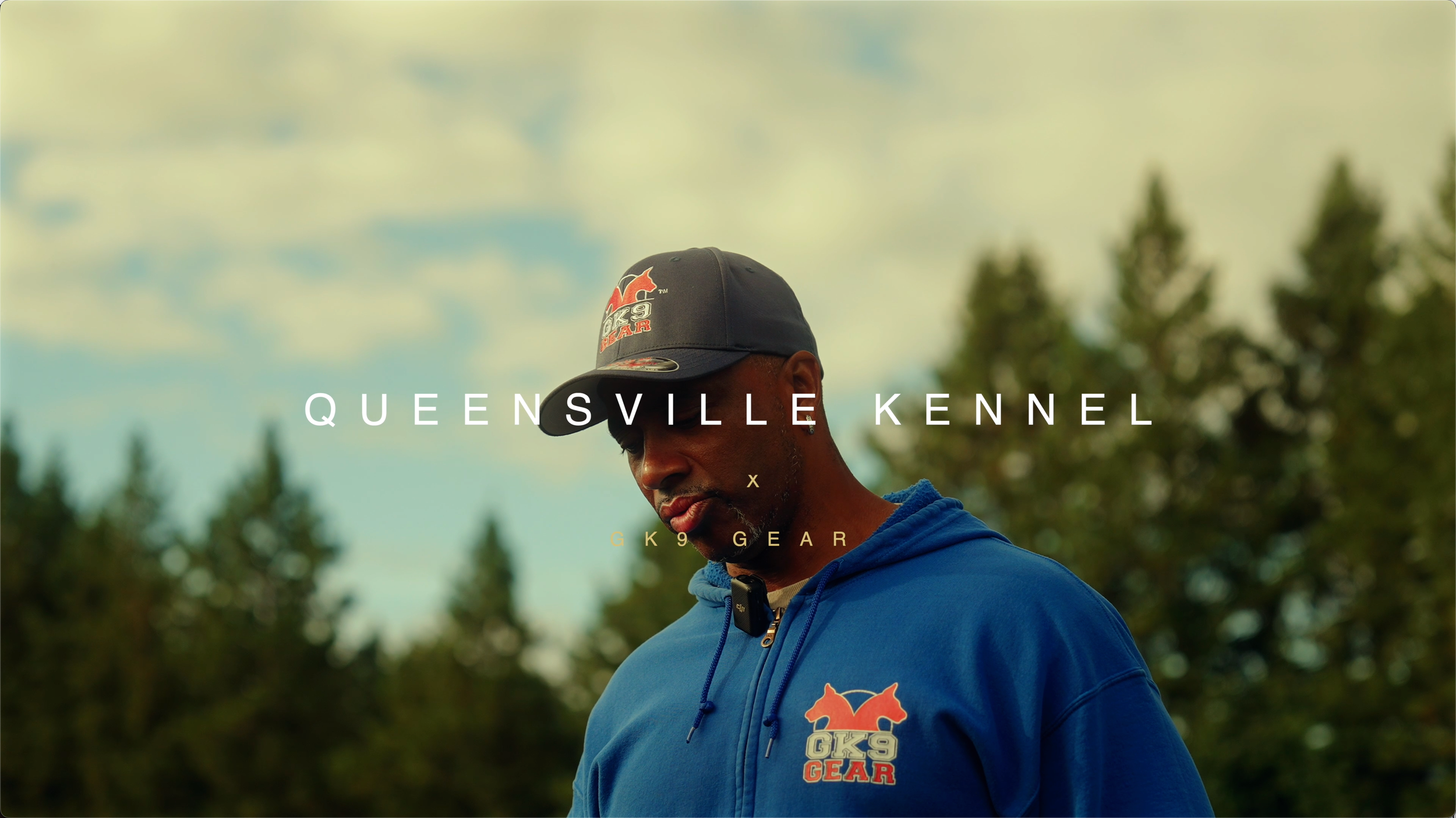 A man wearing a blue hoodie and a black baseball cap with a logo, standing outdoors with trees and a cloudy sky in the background.