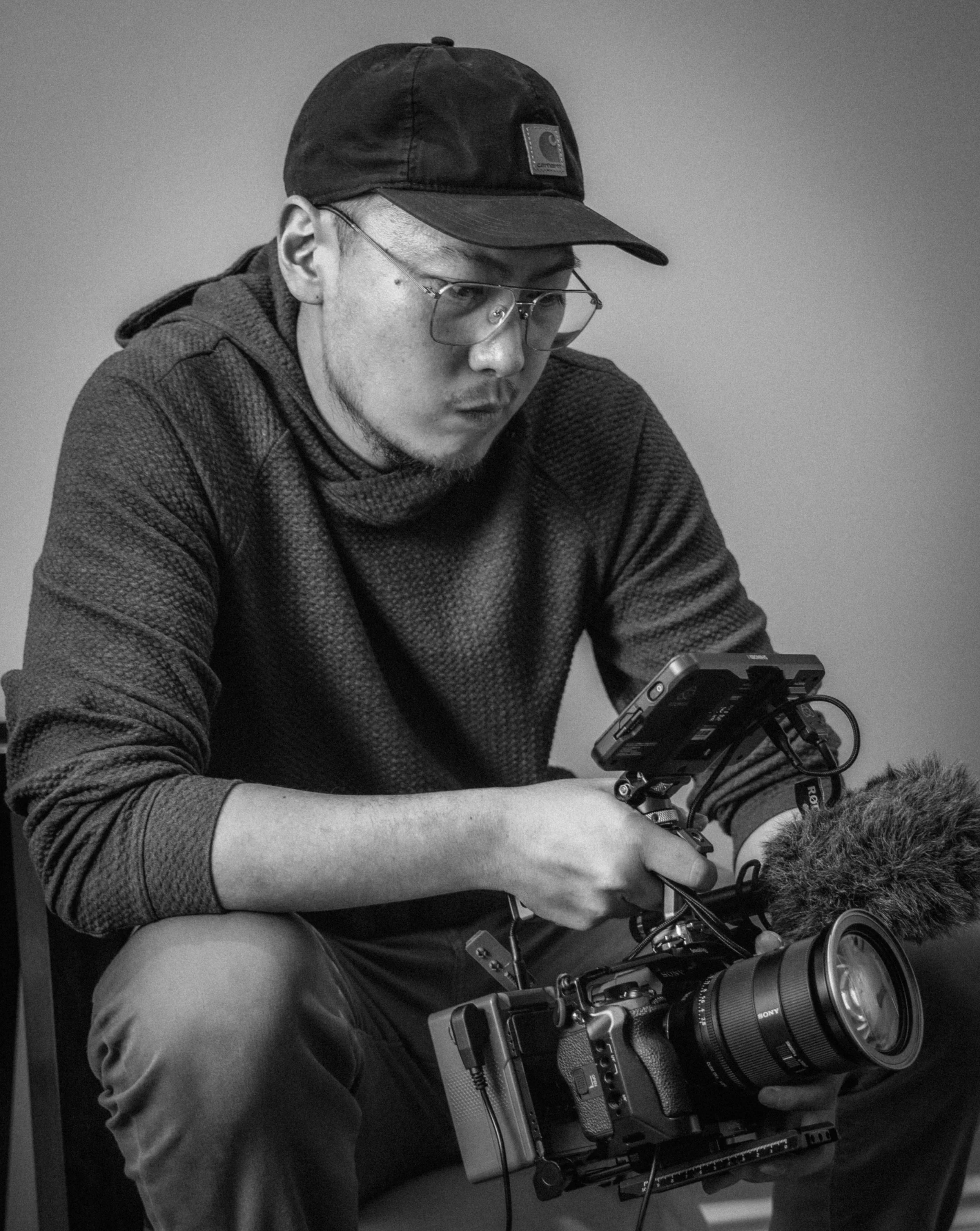 A man wearing a baseball cap, glasses, and a textured sweater, operating a professional video camera with a microphone attached.