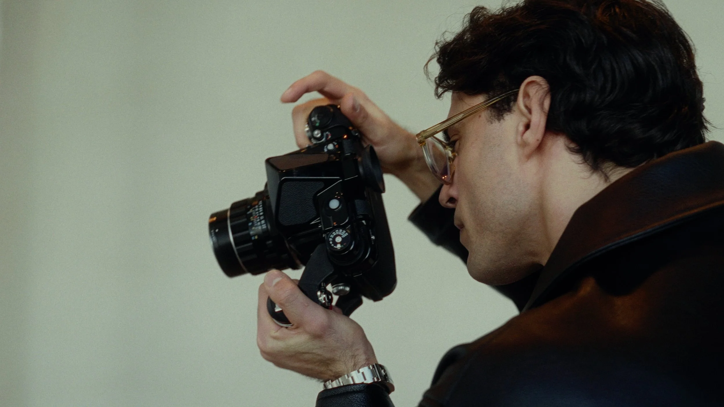 A man with curly dark hair and wearing glasses is looking through the viewfinder of a vintage camera, holding it with both hands, with a plain light-colored background.