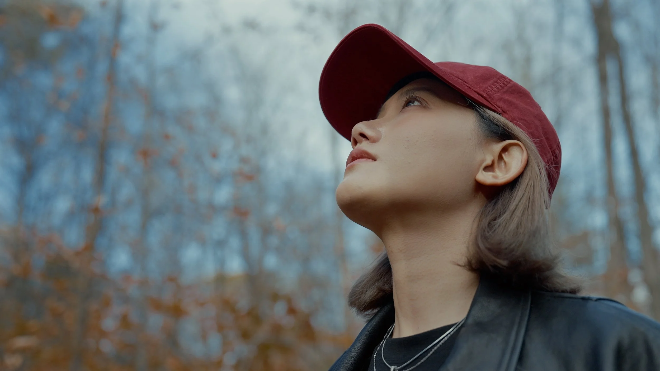 Side profile of a young woman wearing a red cap outdoors in a wooded area with bare trees in autumn, looking upward.