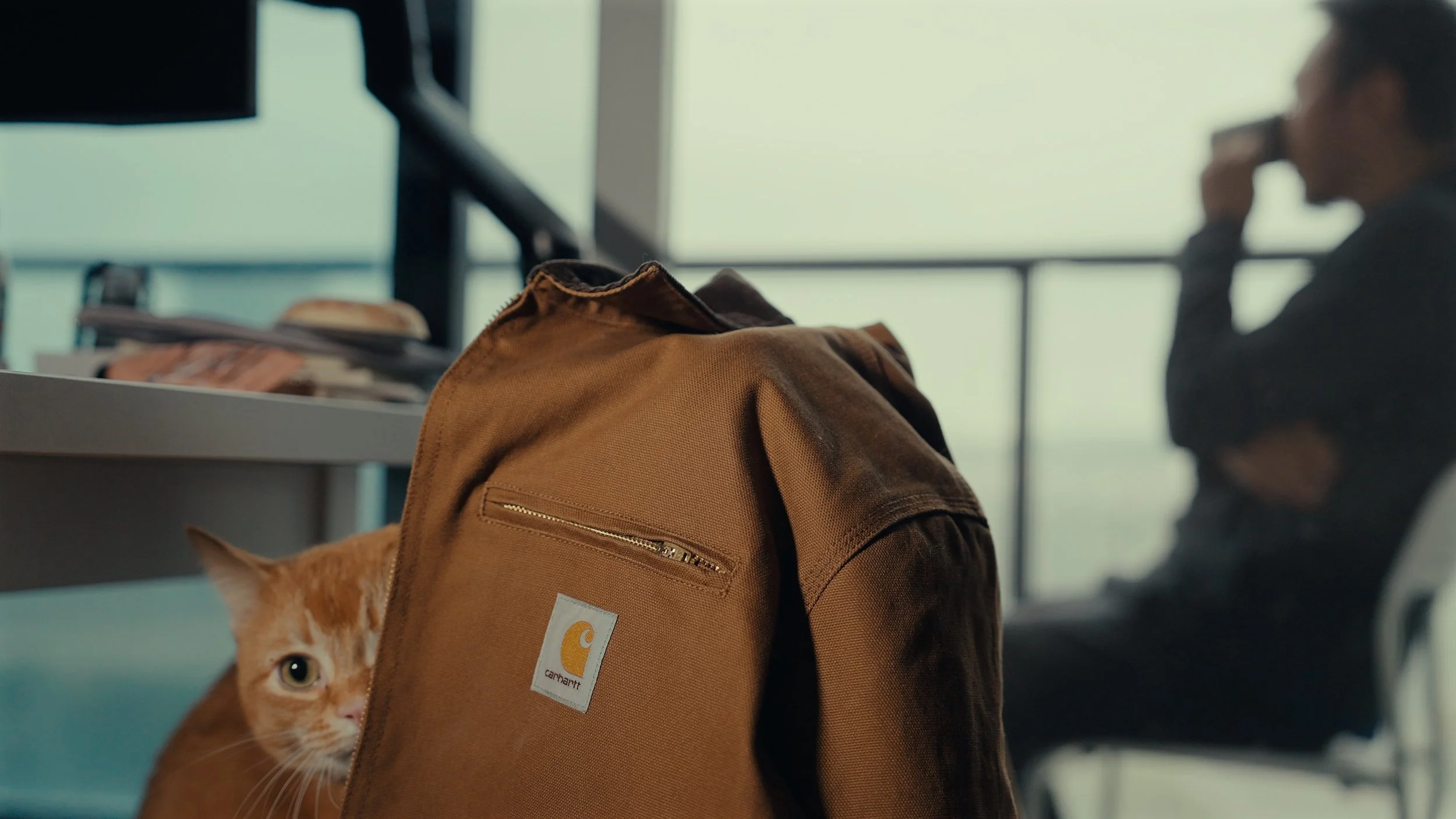 An orange tabby cat peeking out from behind a brown Carhartt backpack on a table, with a woman sitting in the background talking on her phone in an airport or waiting area.