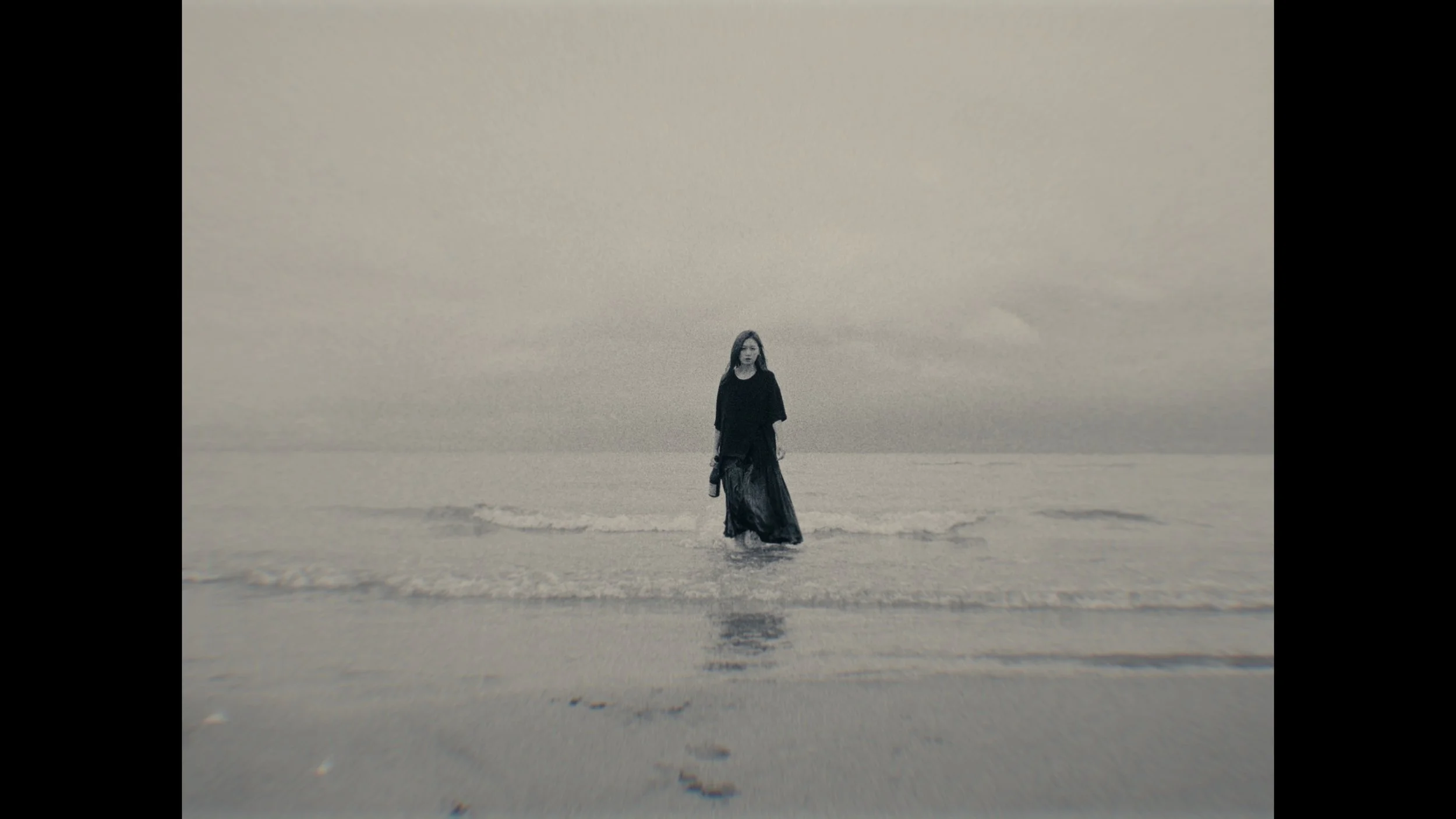 A woman in a dark dress walks in shallow water on a beach, with a gray overcast sky in the background.