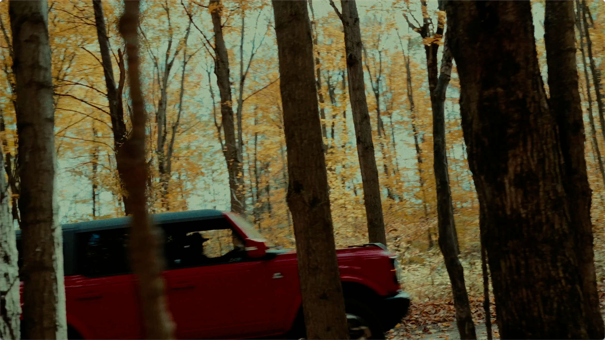A red truck driving through a forest with orange and yellow fall leaves on the trees.