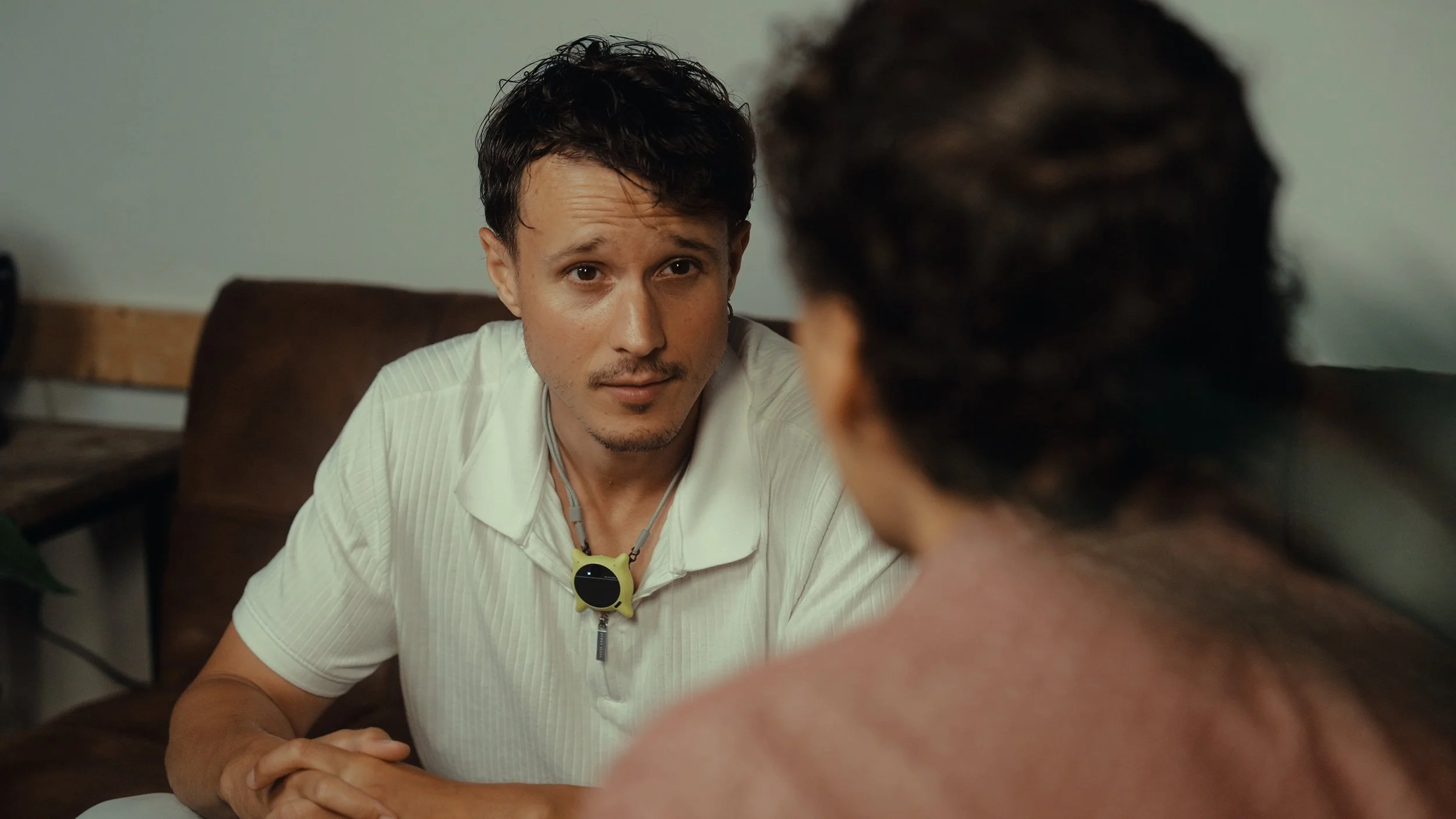 A man with short dark hair, wearing a white shirt and a clip-on yellow and black device around his neck, is sitting at a table and looking at another person whose back is to the camera. They are having a conversation in a room with a green wall and b