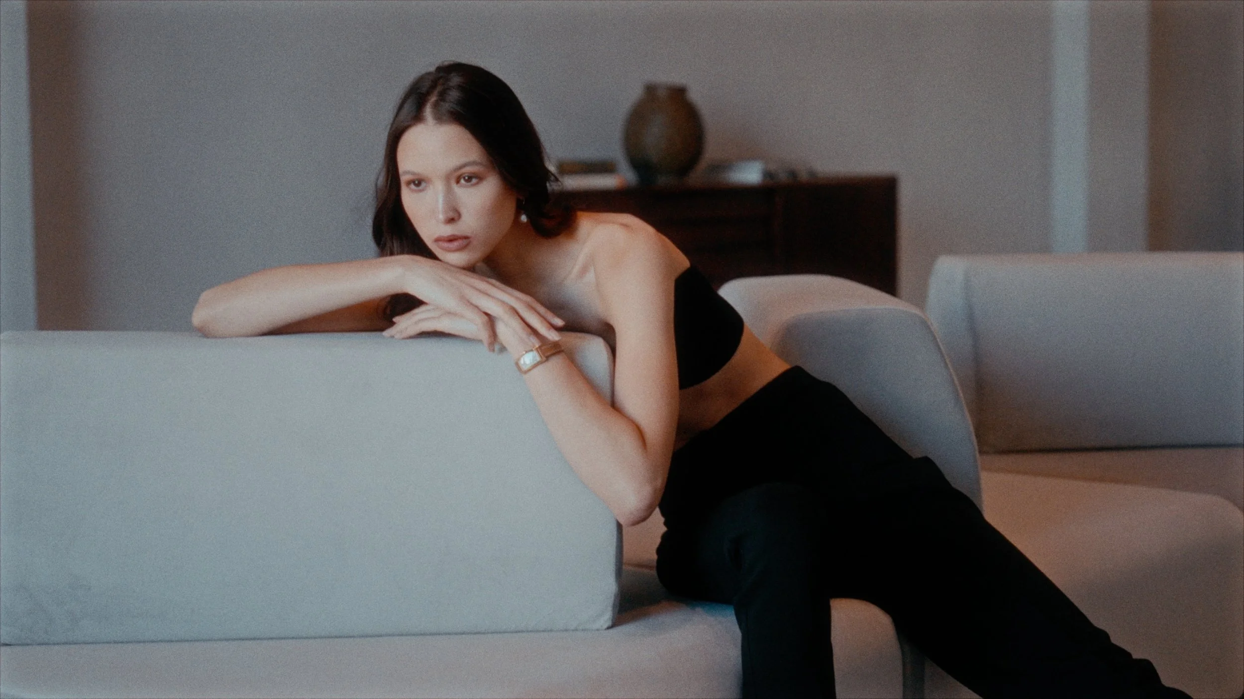 A woman with dark hair and fair skin leaning on a white couch, wearing a black strapless top and black pants, with a watch on her wrist, in a modern living room.