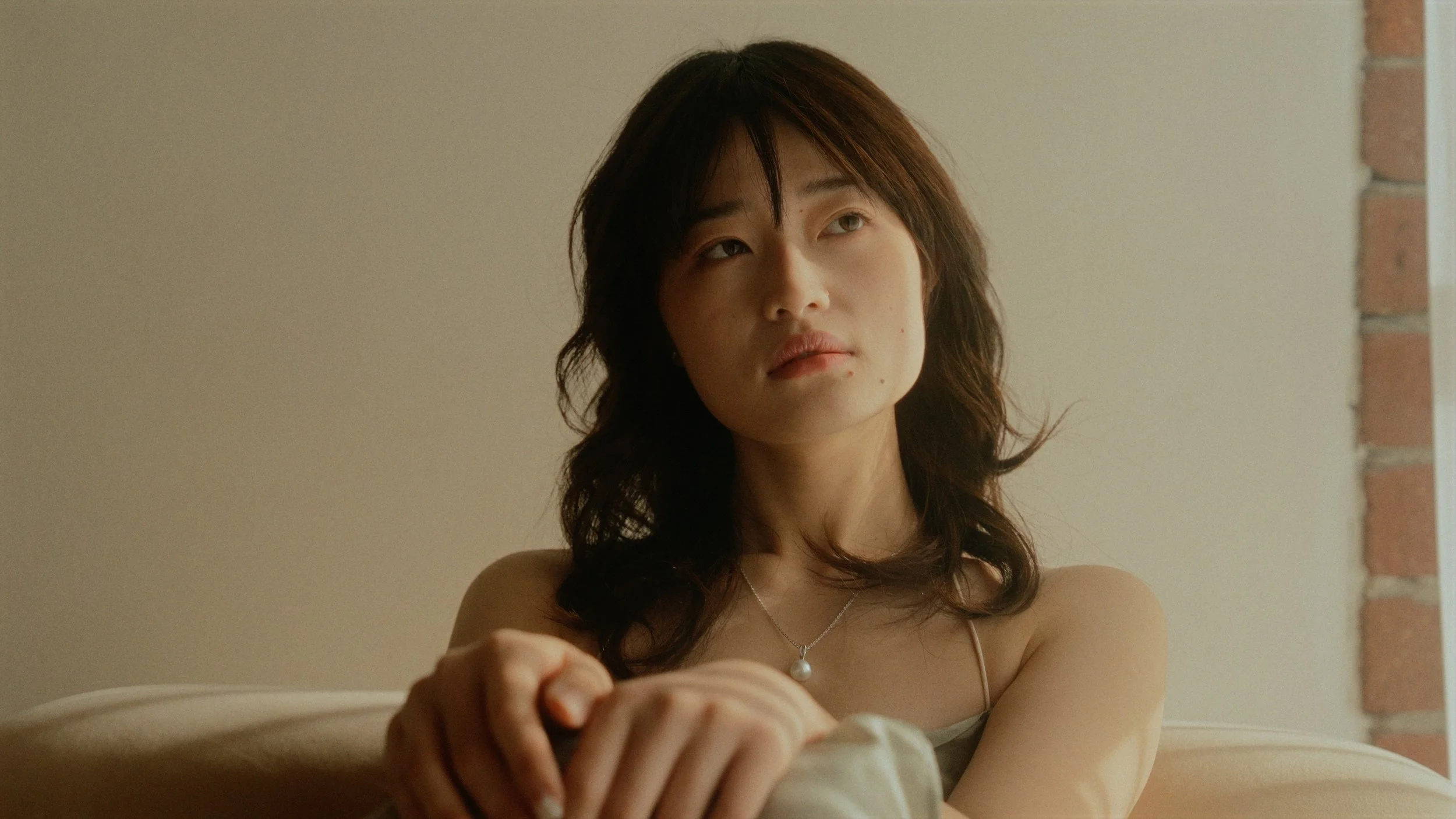 A woman with shoulder-length brown hair and a neutral expression, sitting indoors near a brick wall and looking slightly upward.