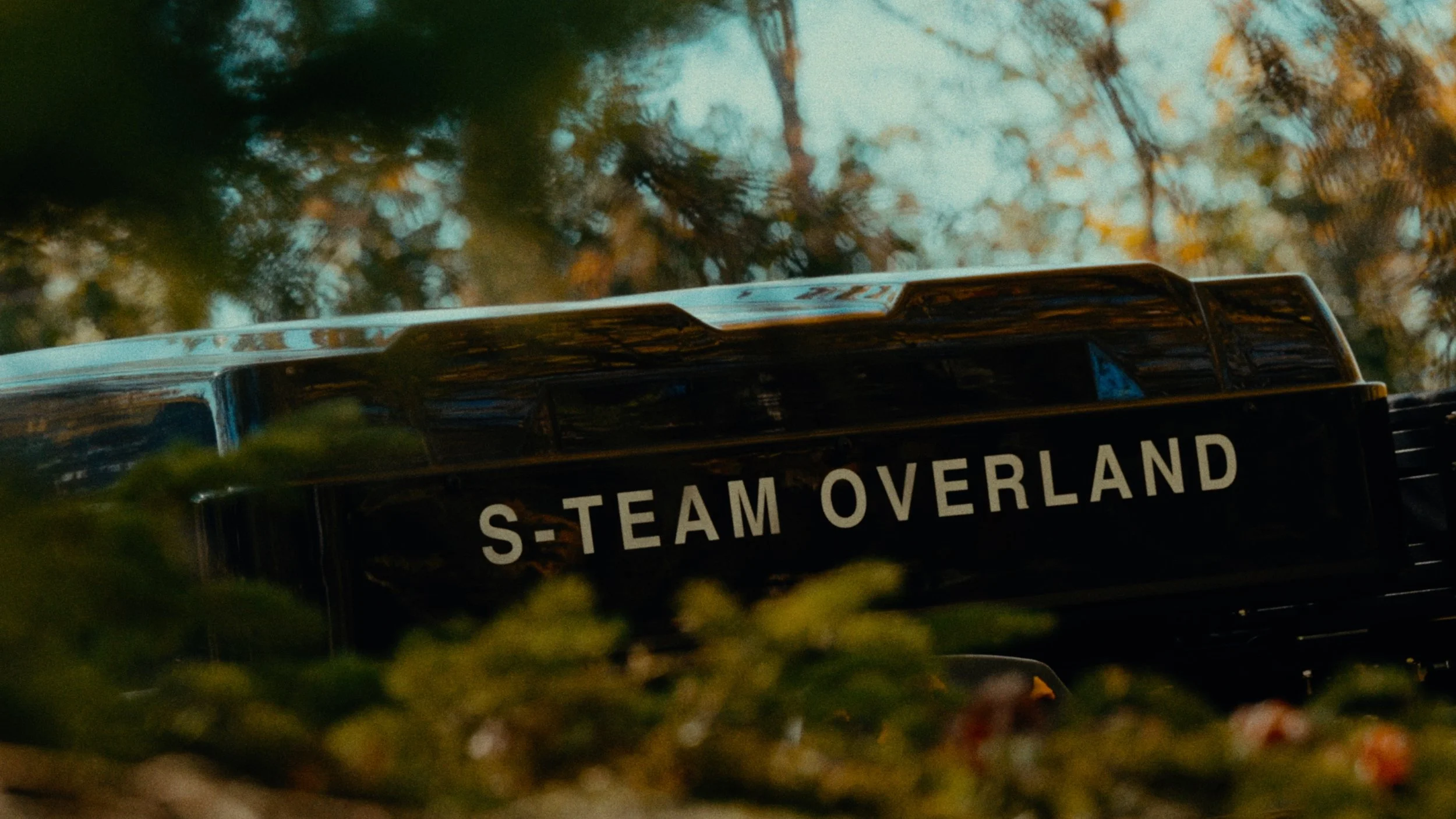 Close-up view of a black vehicle with the text 'S-TEAM OVERLAND' on the side, surrounded by trees and foliage, with blurred greenery in the foreground.