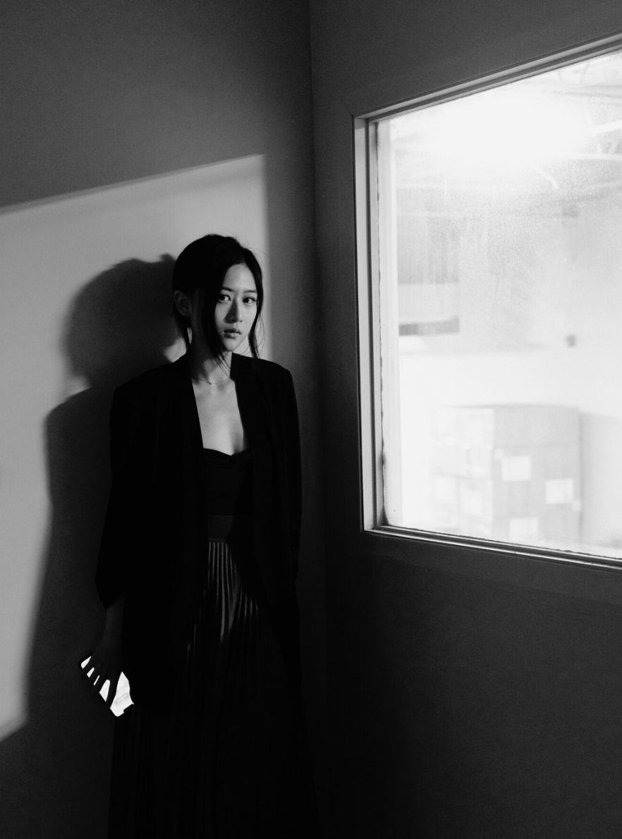 A woman standing indoors in front of a plain wall and a window, dressed in dark clothing, with a serious expression, in black and white photography.