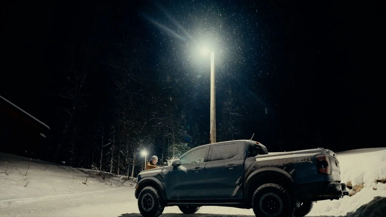 A snowy night scene with a pickup truck parked on the snow-covered ground, illuminated by a streetlight, with a person standing near the truck in the background.