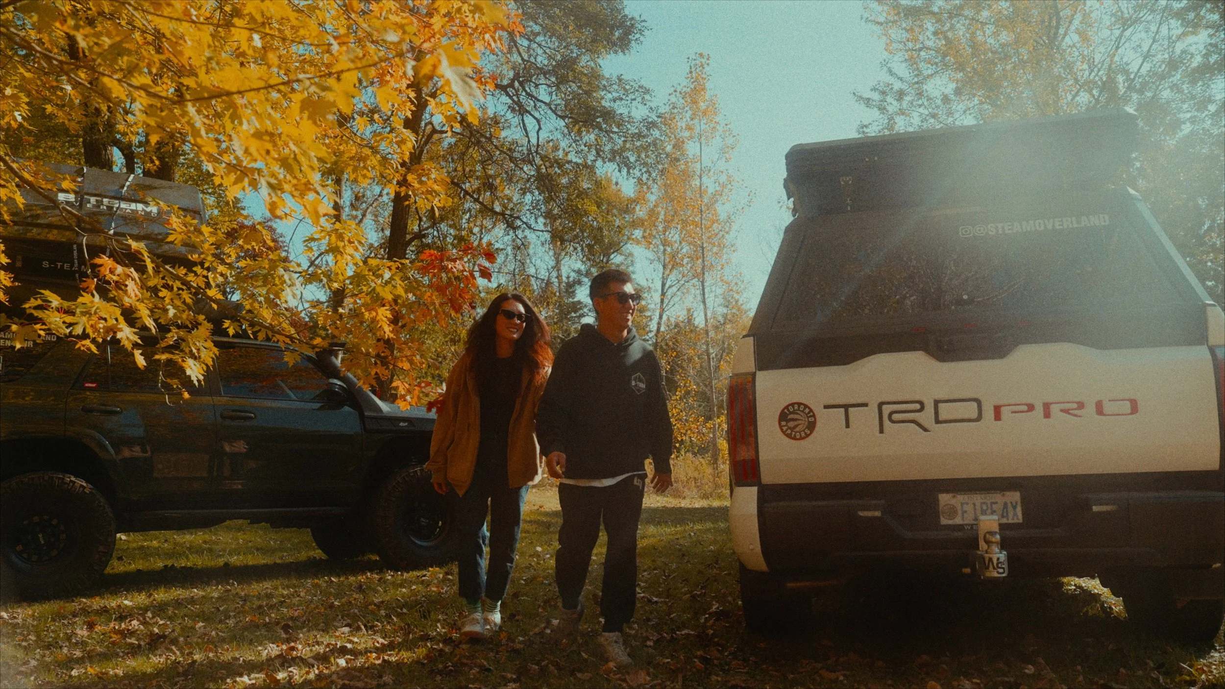 A man and woman walking outdoors on a sunny day, surrounded by trees with autumn-colored leaves, beside a white Toyota TRD Pro SUV and a black off-road vehicle with a roof rack.