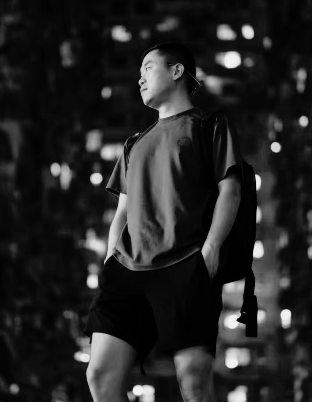 A young man in a casual outfit with a backpack standing outdoors at night, looking to the side with a contemplative expression, illuminated by city lights in the background.