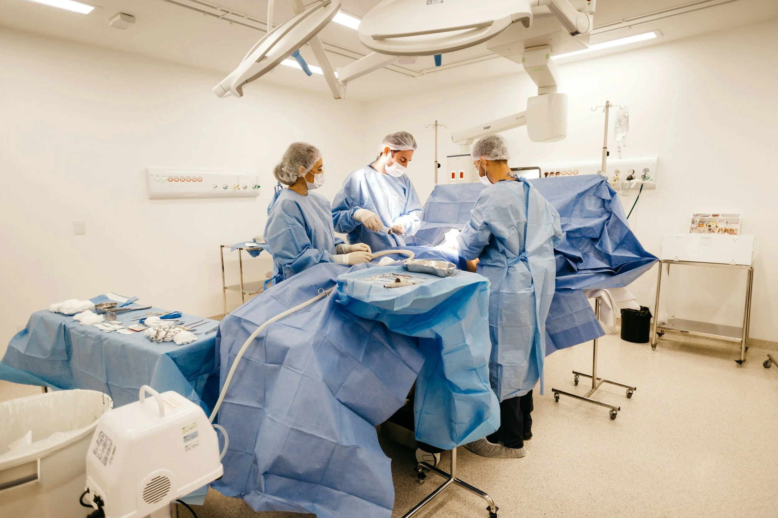Cuatro cirujanos en operación en un quirófano, vestidos con ropa quirúrgica, mascarillas y gorros, realizando una cirugía asistida por instrumentos médicos y equipos de monitoreo.