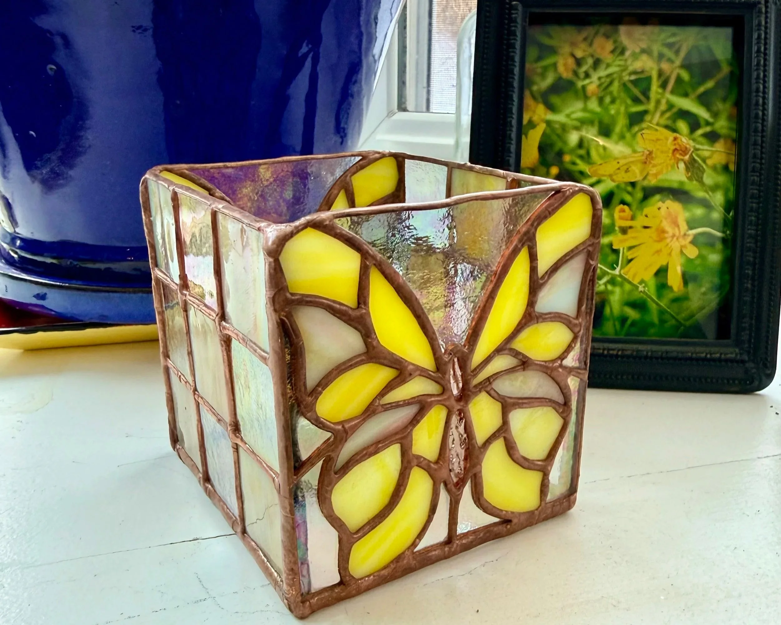 A stained glass butterfly pattern candle holder with yellow and white glass, positioned on a white surface, with framed floral artwork and a blue ceramic object in the background.