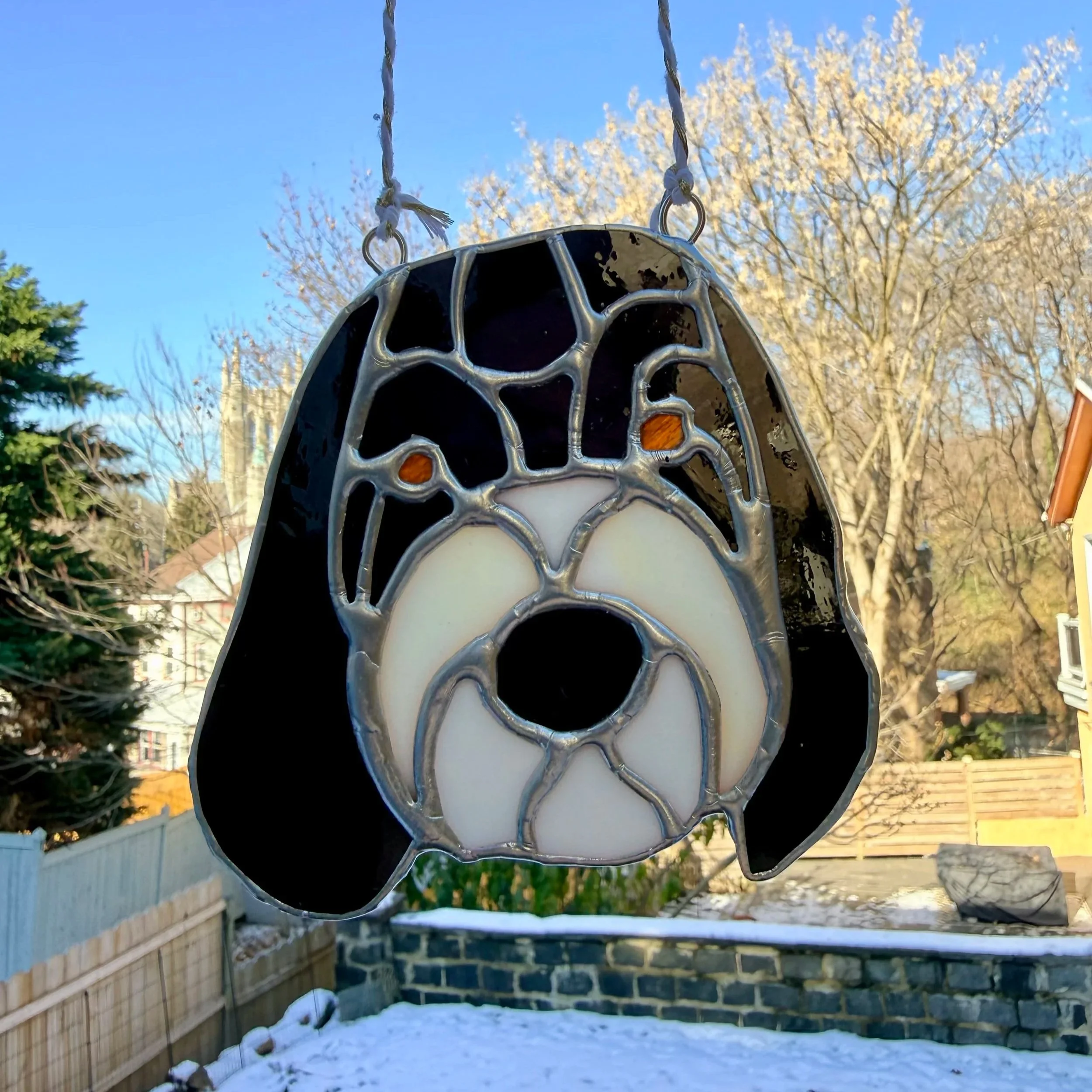 Stained glass ornament shaped like a dog's face with black, white, and brown details, hanging outdoors against a clear sky with bare trees and houses in the background.