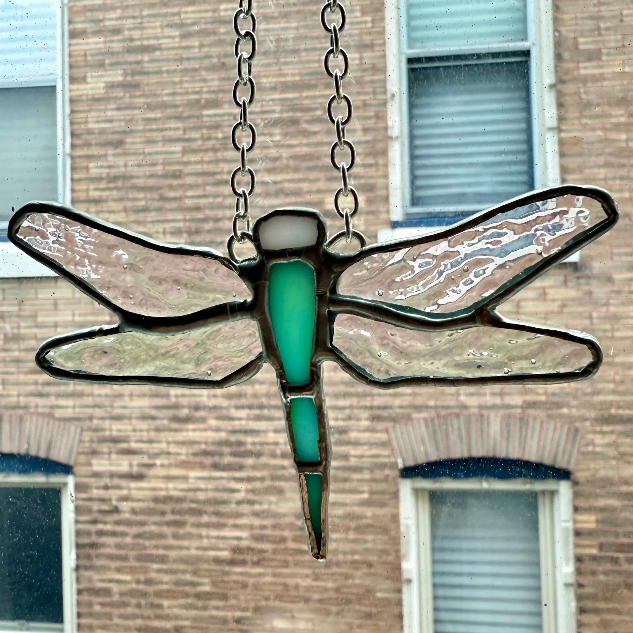 Stained glass dragonfly hanging from a chain outside a window, with a brick building in the background.