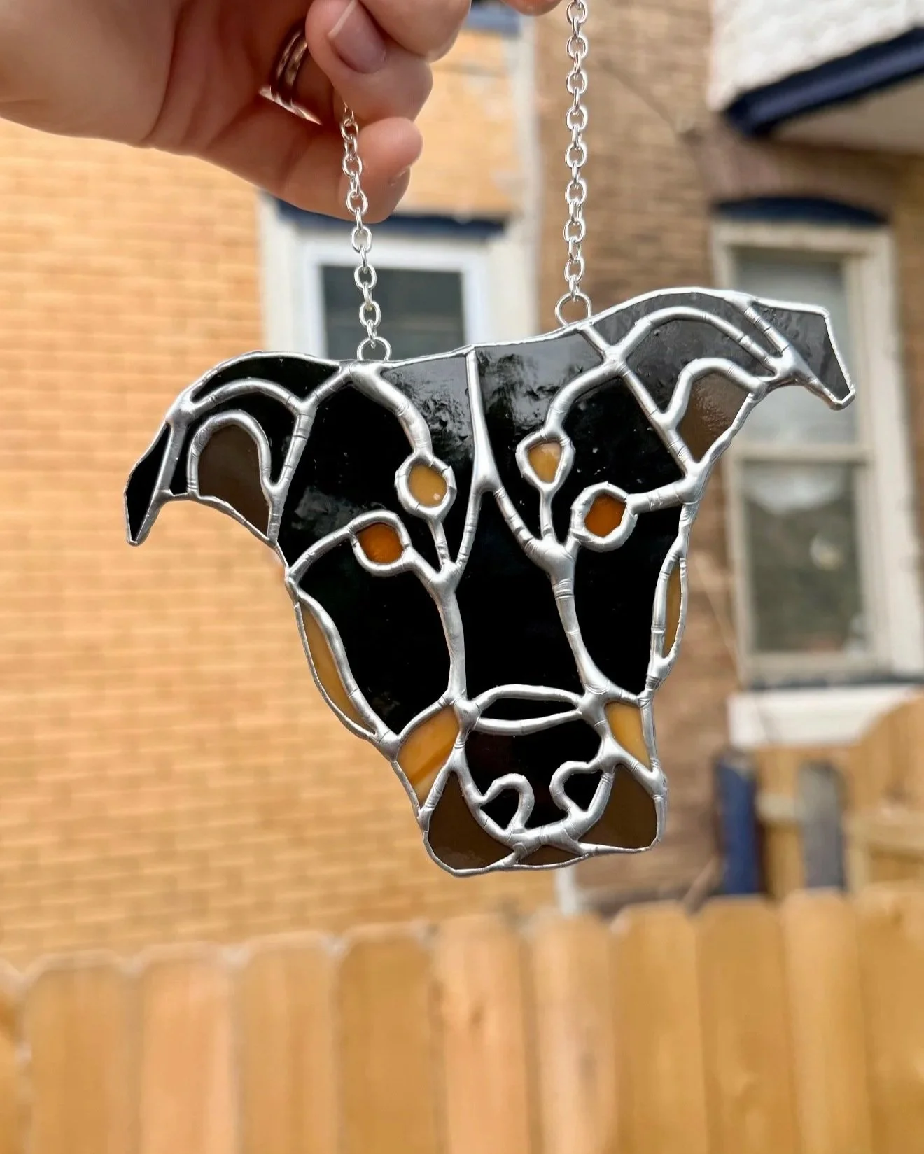 Stained glass ornament of a dog's face, held by a hand outside with a brick house and windows in the background.