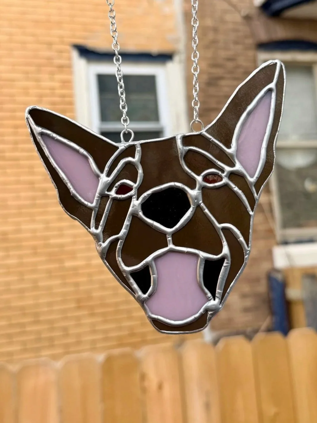 A stained glass suncatcher in the shape of a dog's face, with pink, black, and brown glass pieces, and a chain hanging from the top.