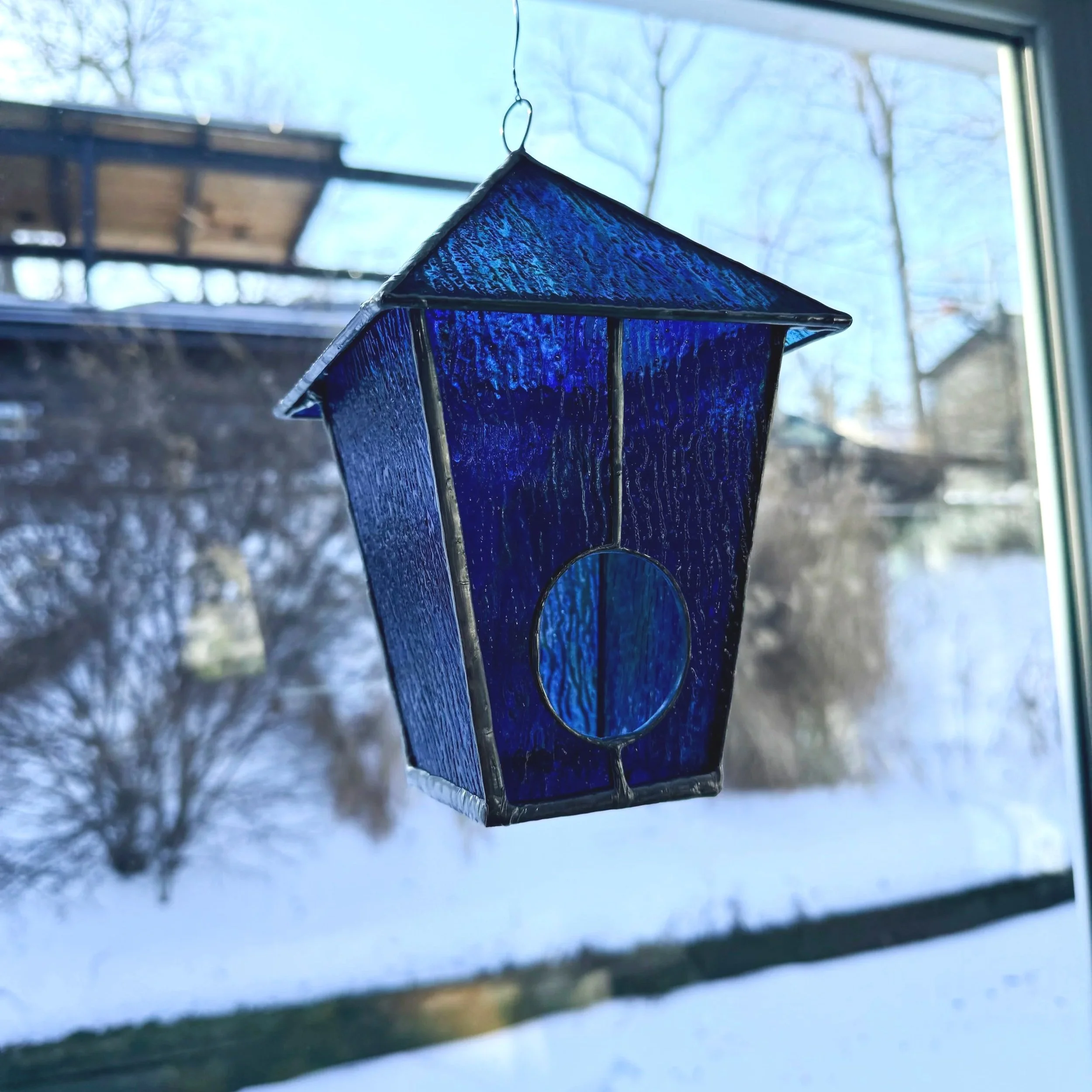 Blue stained glass bird house hanging by a chain in front of a window with a snowy outdoor scene with trees.