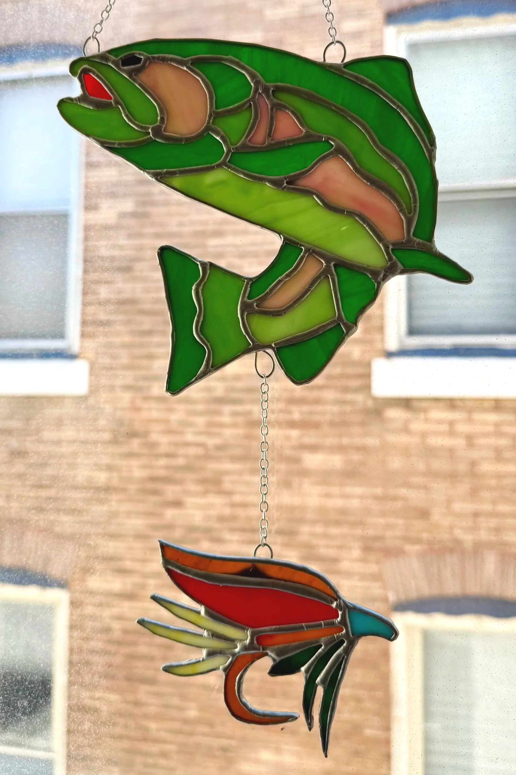 Colorful stained glass hanging of a green fish with a red mouth above a red and orange bird with green wings, hanging on a chain in front of a window.