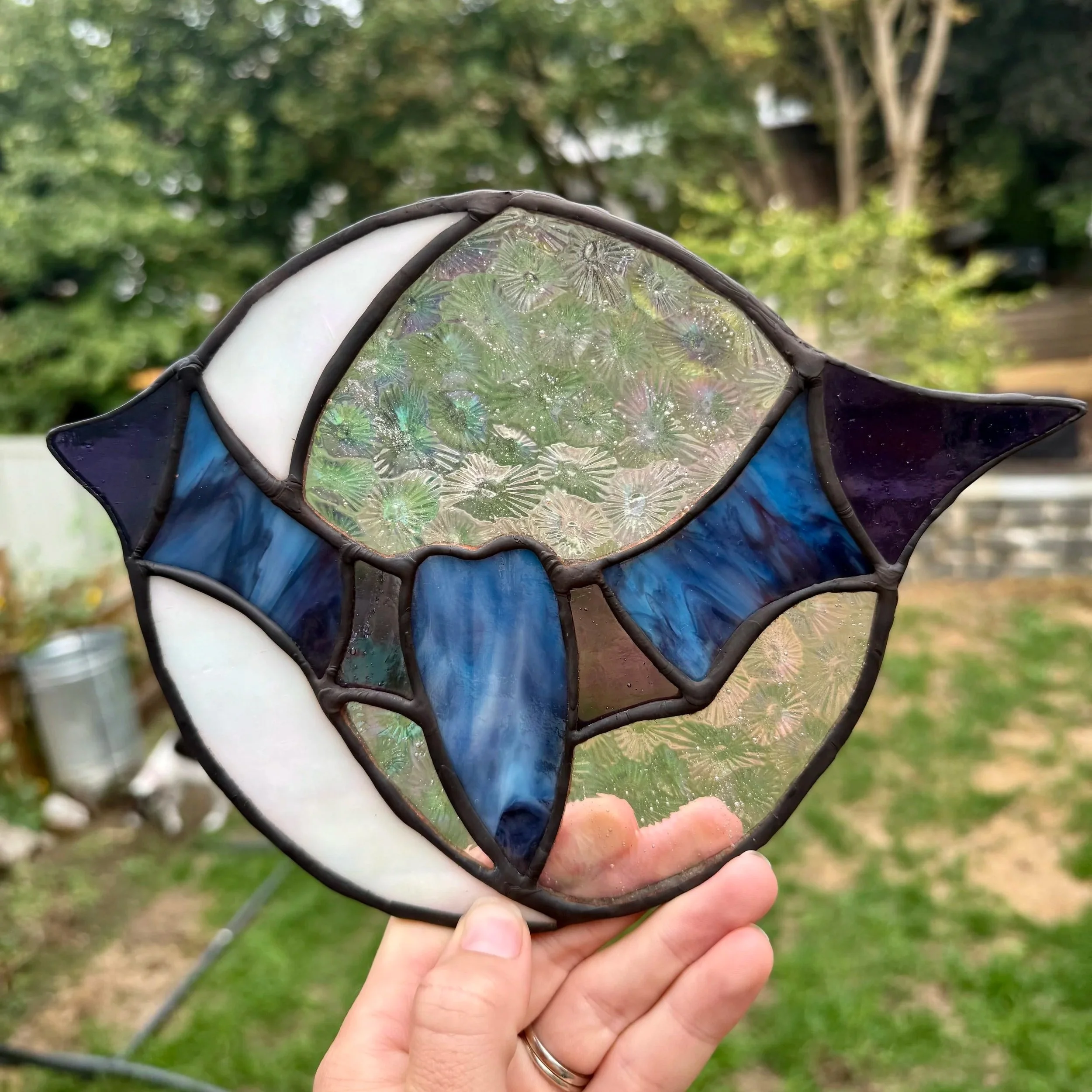 Stained glass art piece with an abstract fish design, featuring blue, white, and iridescent glass sections, held outdoors with trees in the background.