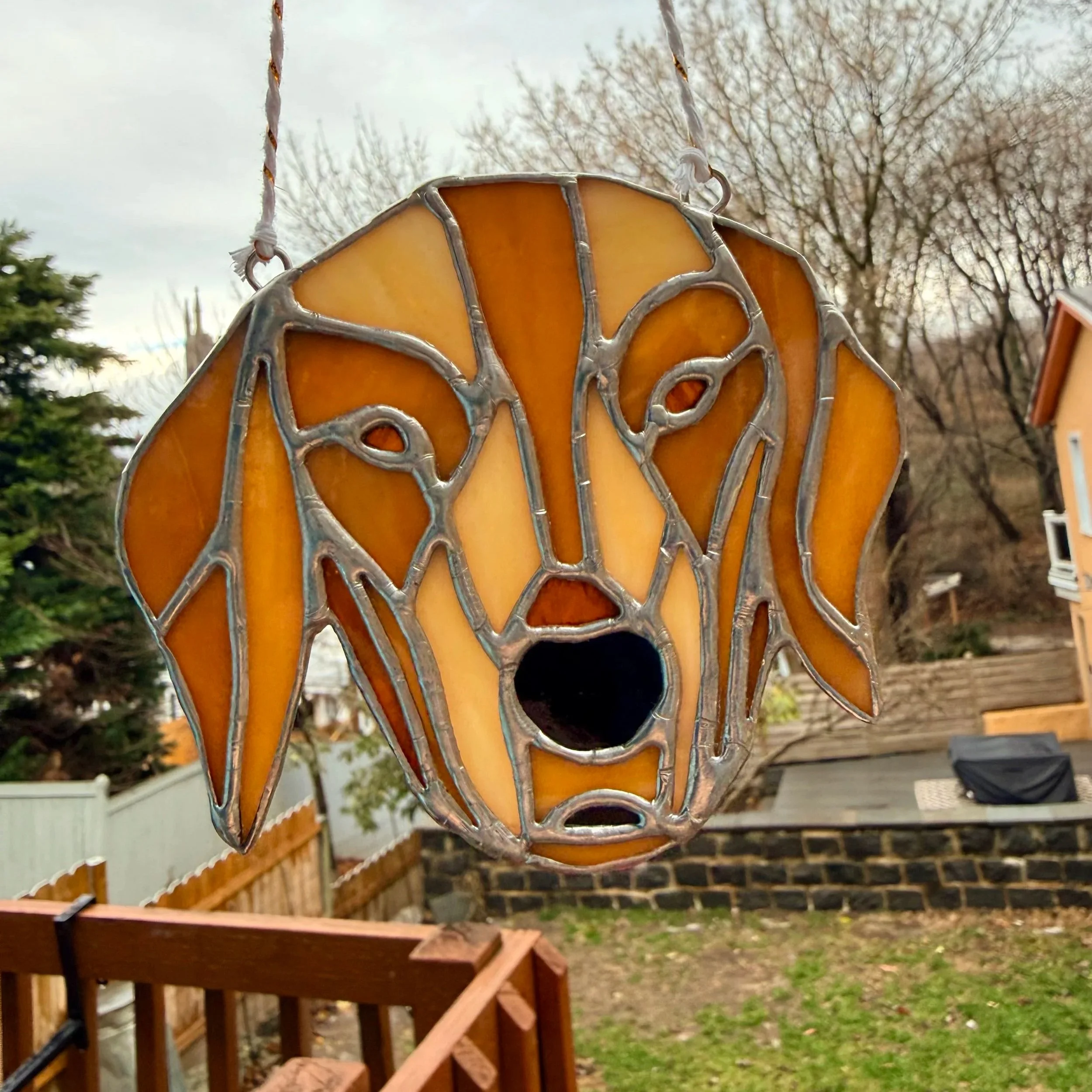 Stained glass garden ornament in the shape of a dog's face hanging outdoors with trees and a house in the background.