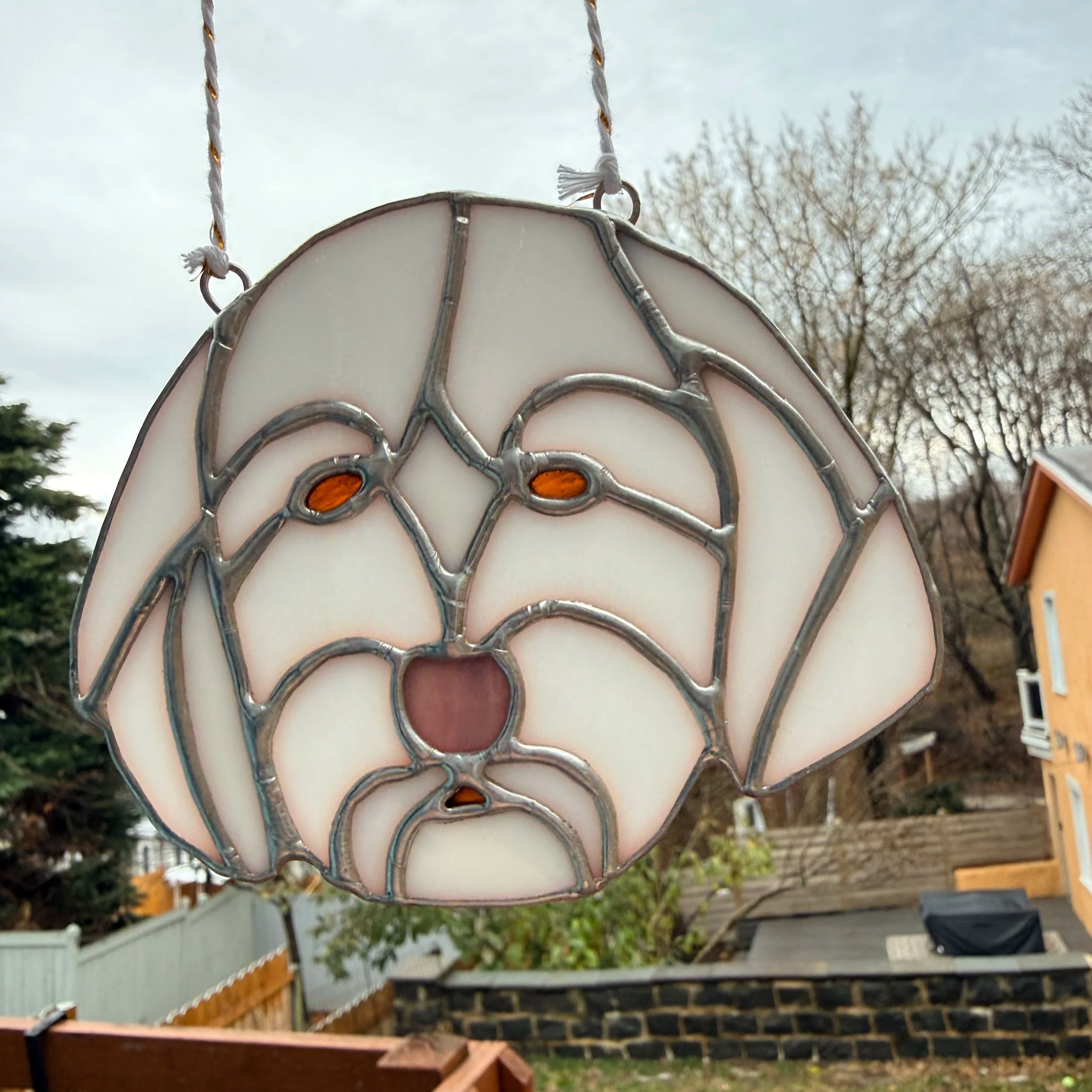A stained glass hanging ornament shaped like a dog's face with tan, black, and pink colors, mounted outdoors with trees and houses in the background.