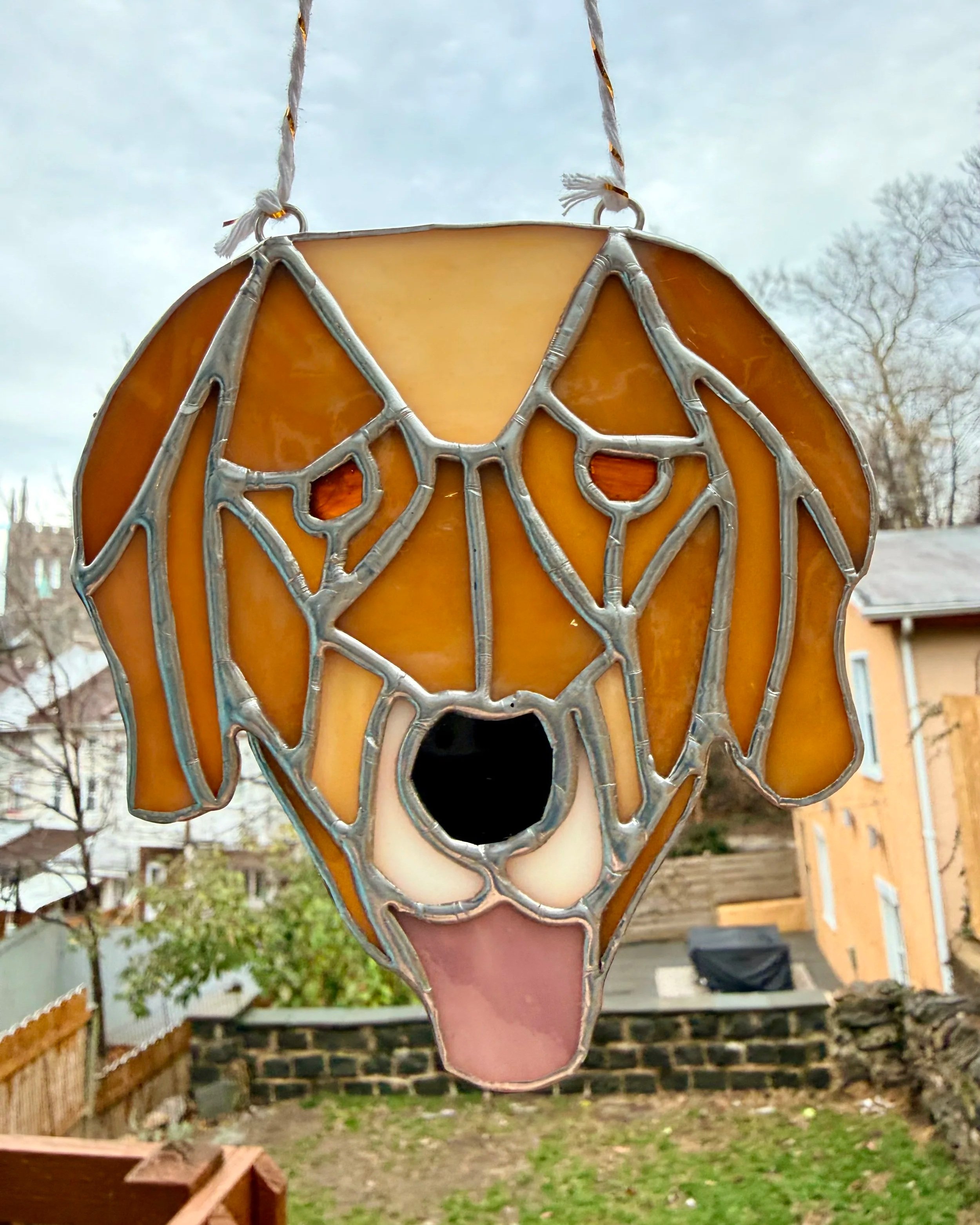 Stained glass hanging ornament shaped like a dog's face with yellow, white, pink and black glass pieces, with a cloudy sky and backyard in the background.