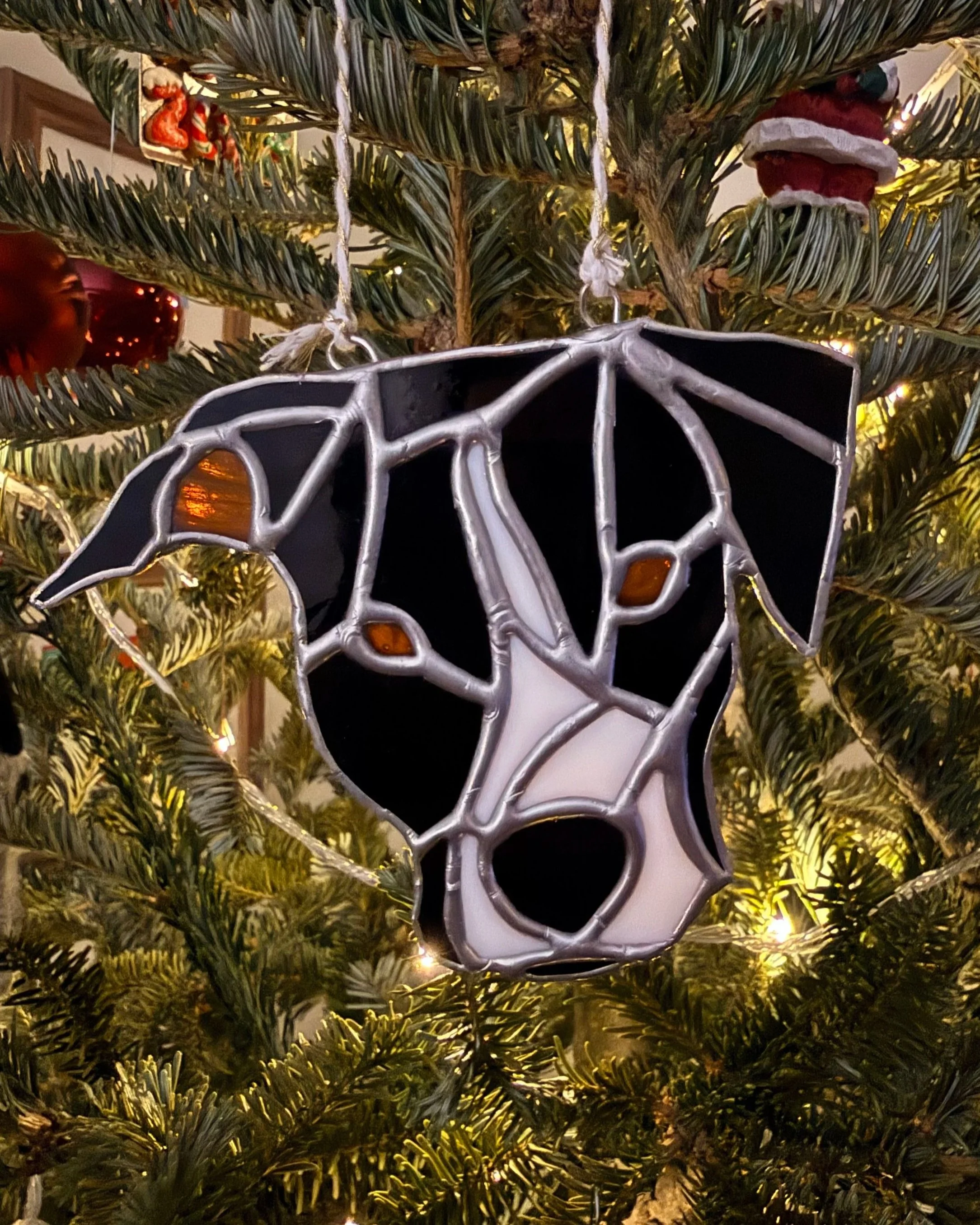 A glittery ornament of a dog's face hanging on a decorated Christmas tree with lights and other ornaments.