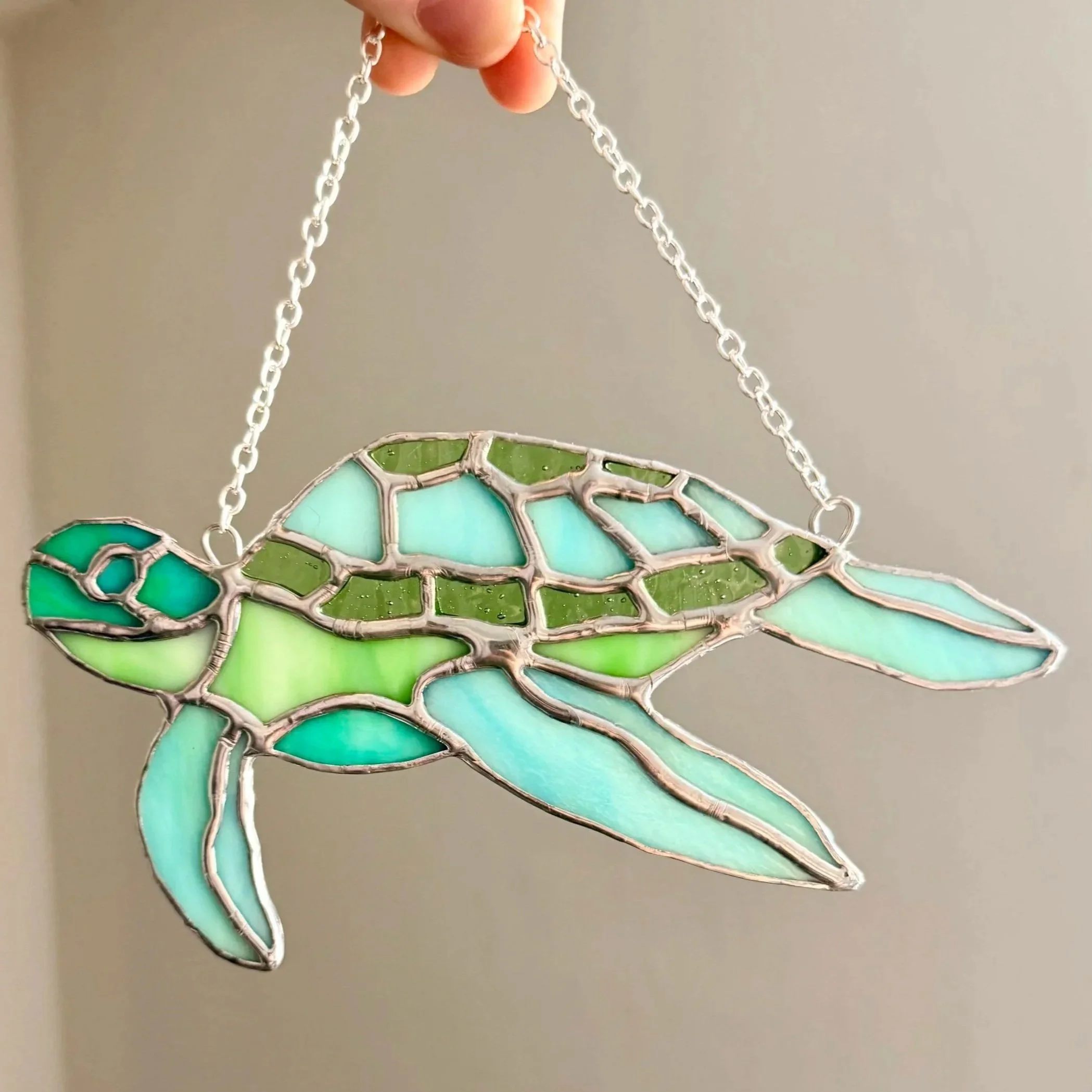 Stained glass hanging ornament in the shape of a dragonfly with shades of green and blue, held by a silver chain.