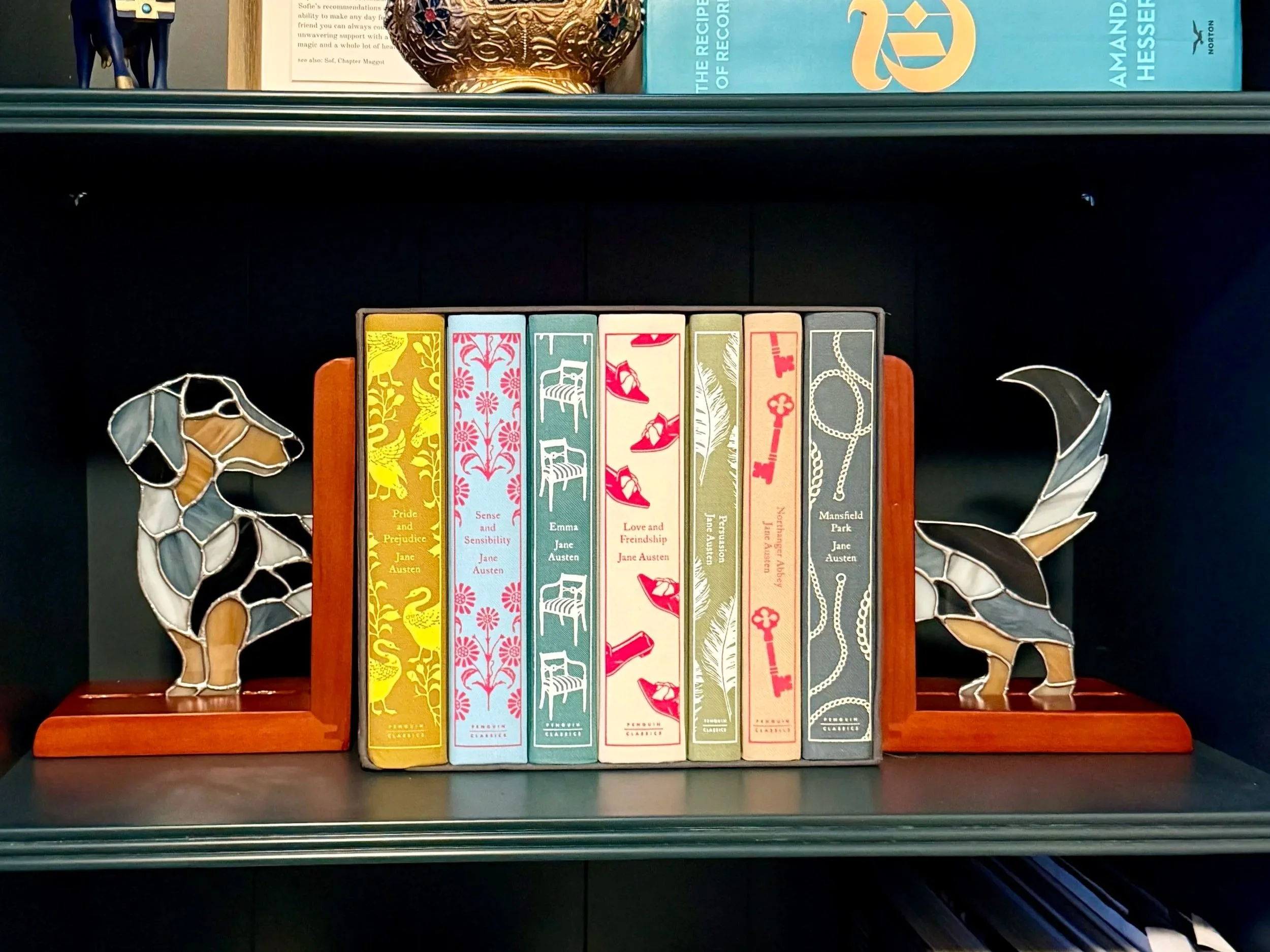 Decorative bookshelf with stained glass dachshund bookends, and a row of books by Jane Austen in colorful covers.