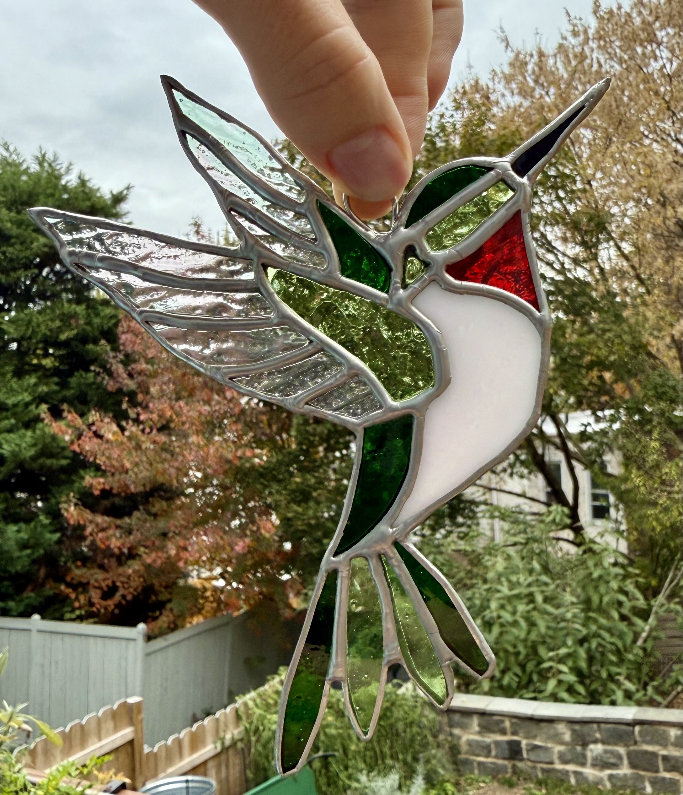 Stained glass hummingbird ornament held up by a hand outdoors, with trees and a wooden fence in the background.