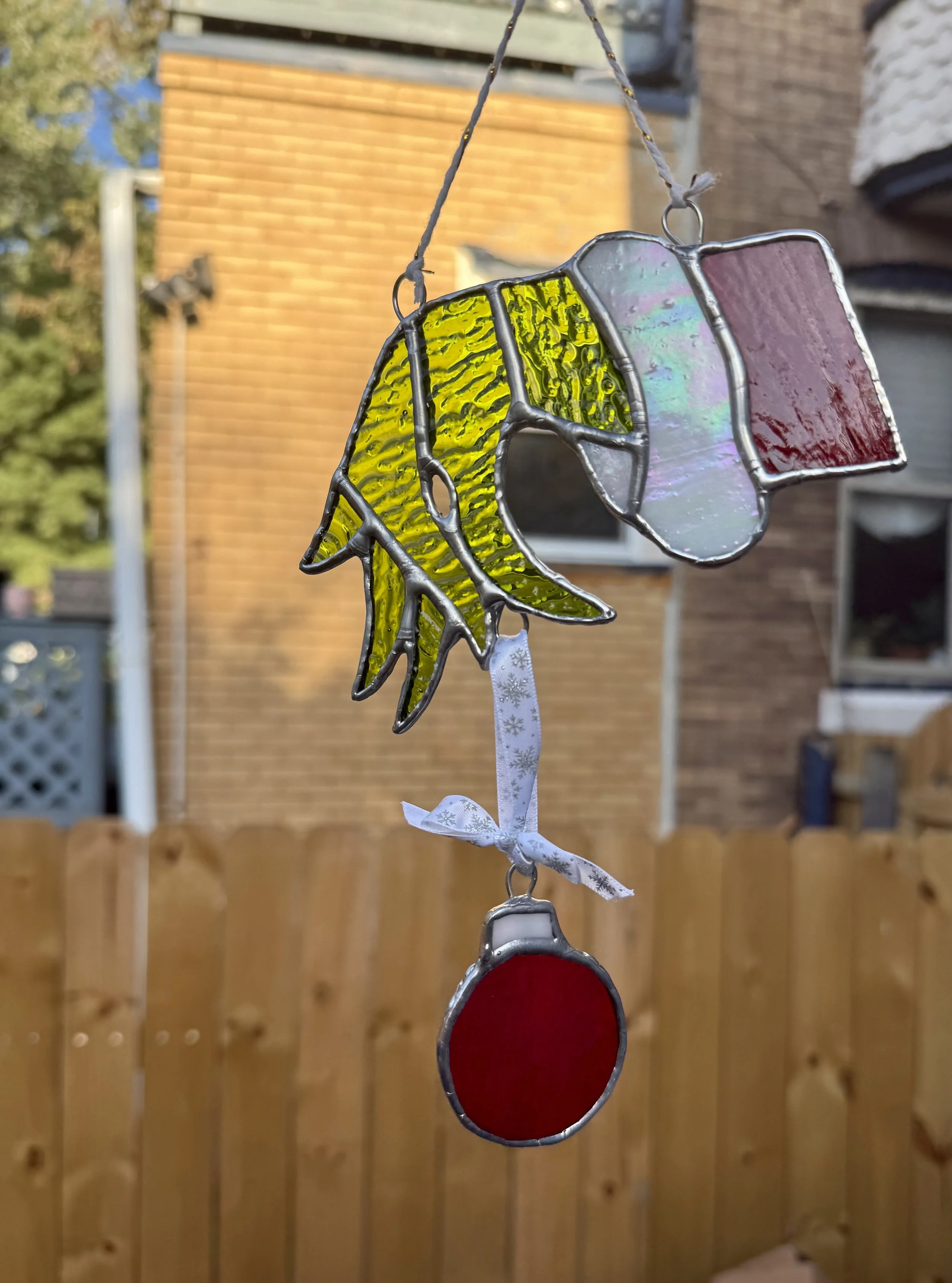 Stained glass Grinch hand with ornament hanging outdoors with houses and trees in the background.