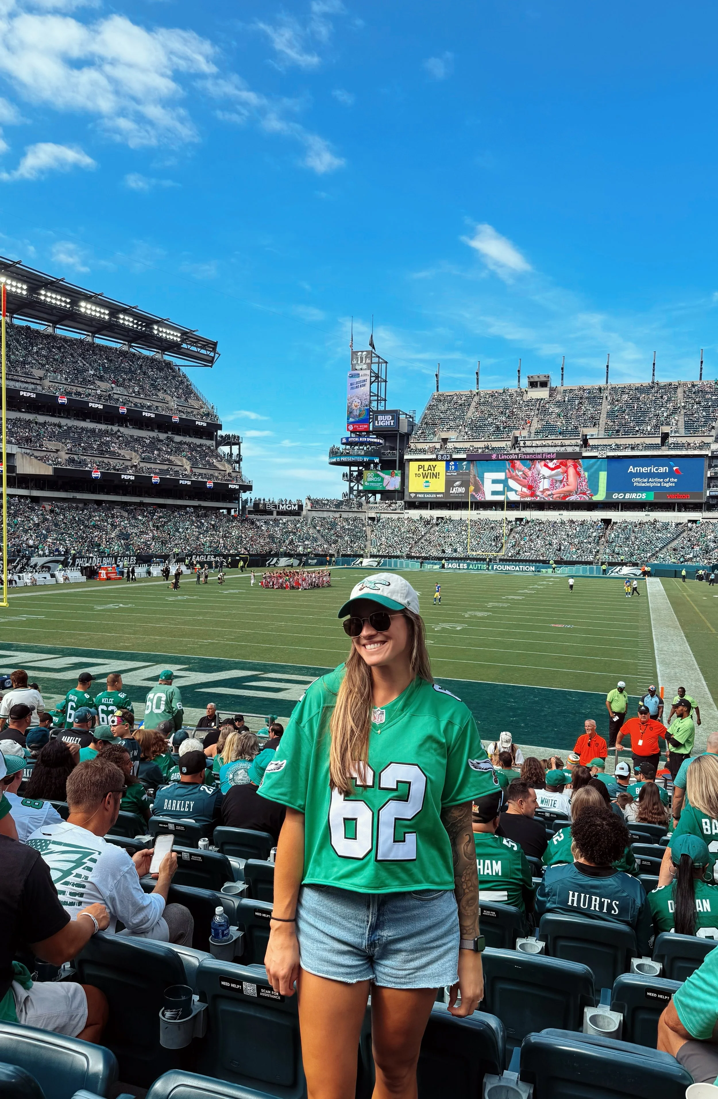 A woman wearing a green Philadelphia Eagles jersey, sunglasses, and a white cap stands smiling at a football game at Lincoln Financial Field. The stadium is filled with fans, and the field is visible in the background under a blue sky.